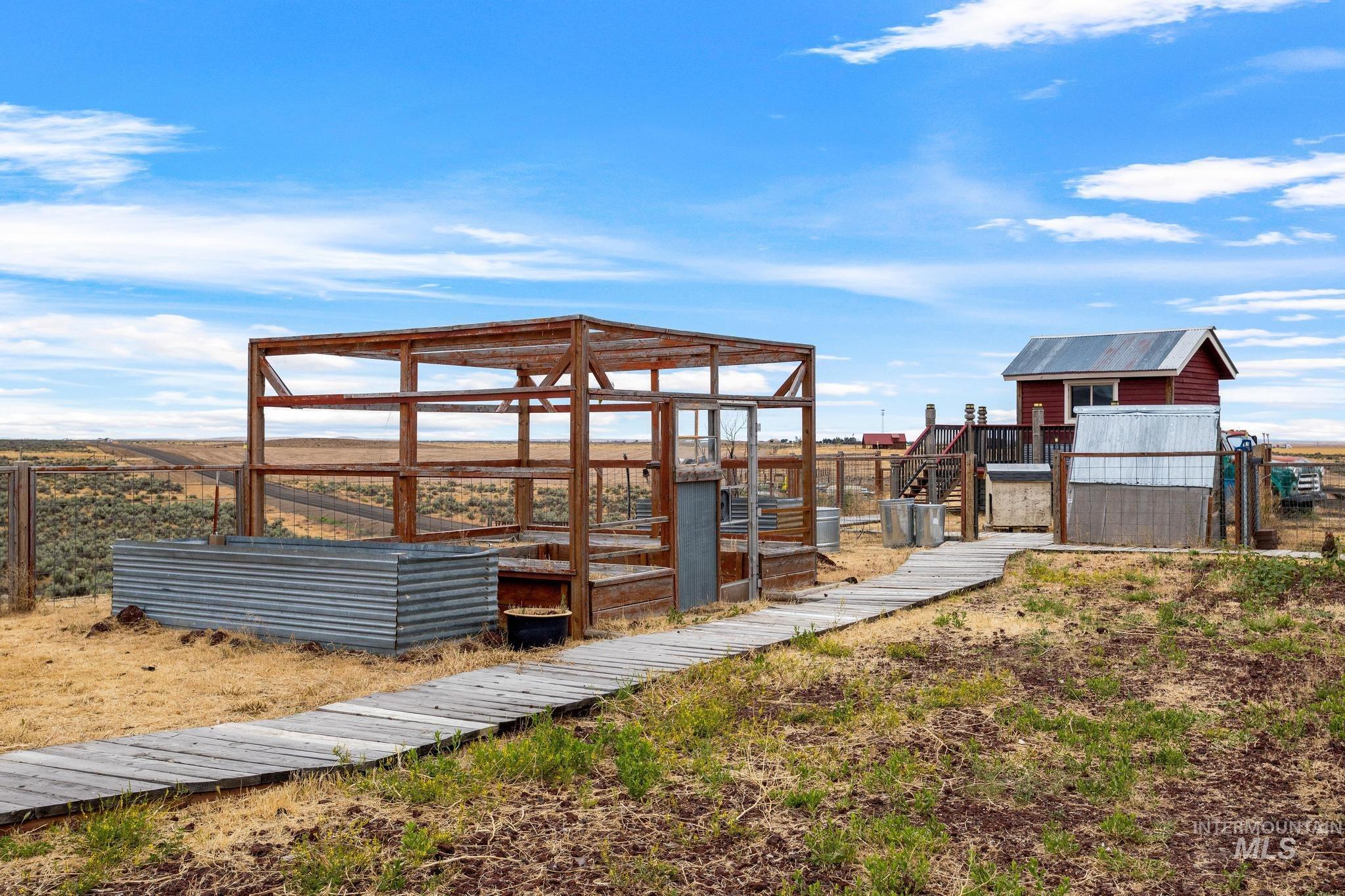 Outdoor wood-framed structure with a corrugated metal planter box, a wooden pathway, and a red-sided outbuilding featuring a metal roof