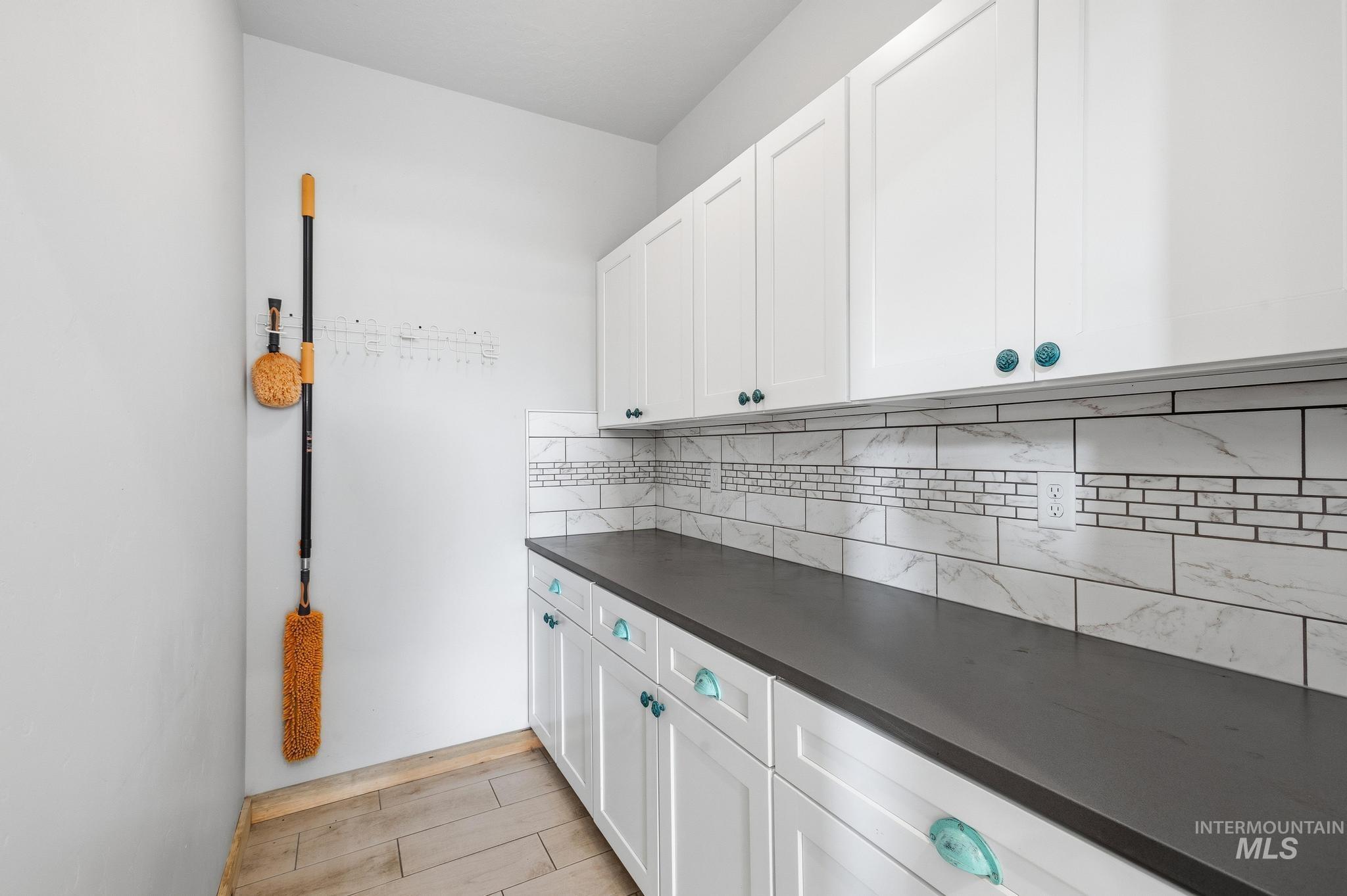Spacious utility area featuring white shaker cabinetry, dark countertops, a subway tile backsplash with mosaic accents, and wood-finish flooring