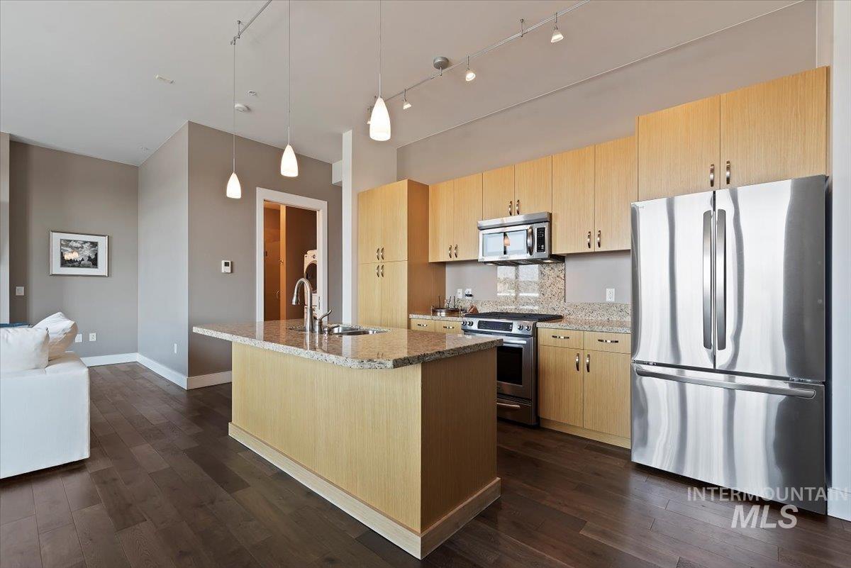 Kitchen with appliances with stainless steel finishes, light brown cabinets, hanging light fixtures, light stone countertops, and dark wood-style floors