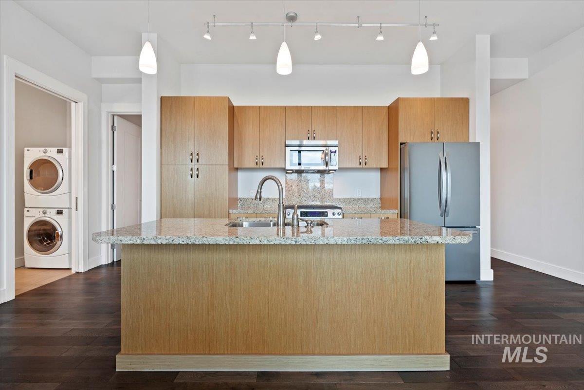 Kitchen with light stone countertops, stainless steel appliances, decorative light fixtures, a center island with sink, and dark wood-type flooring