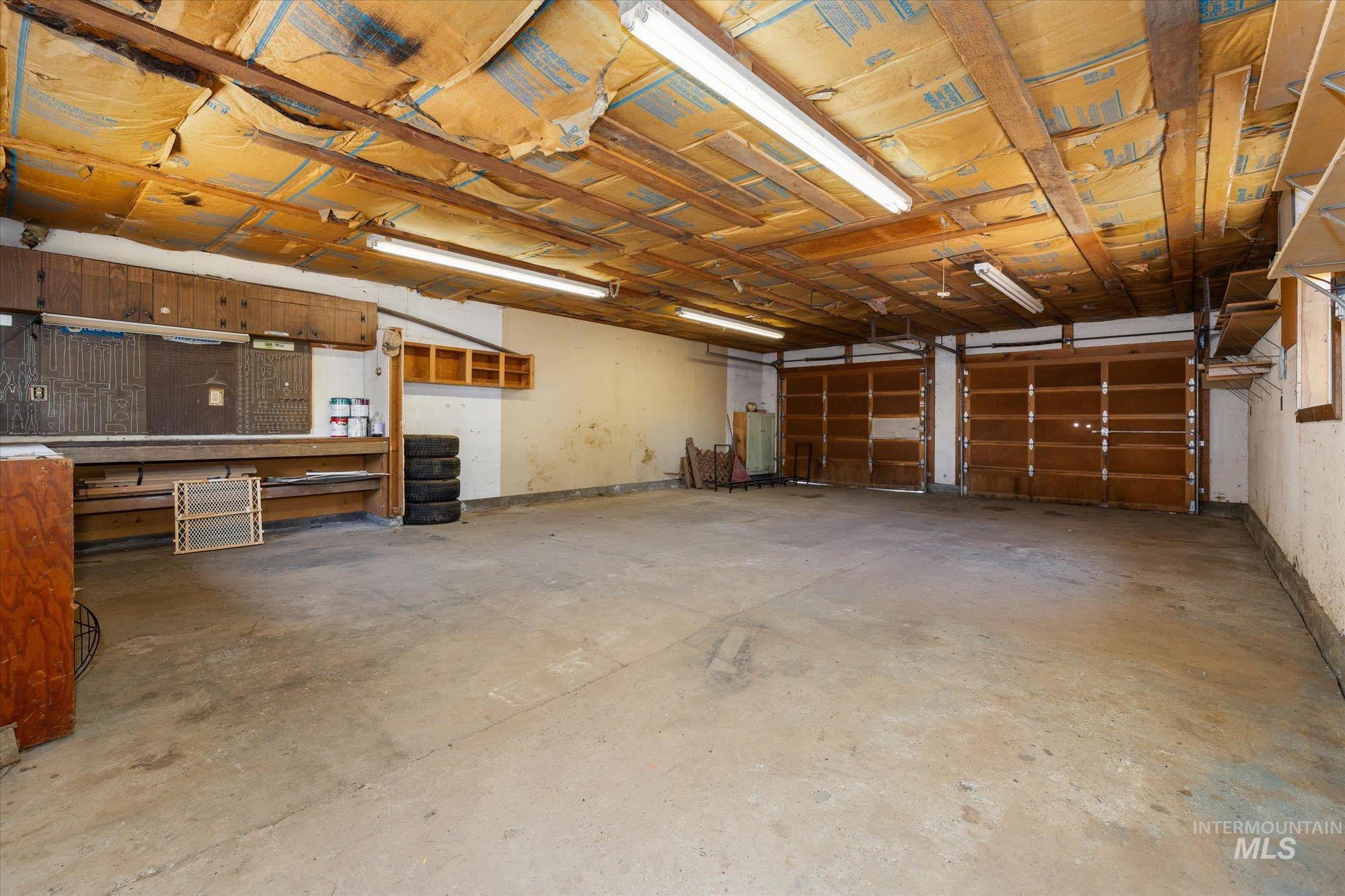 Two-car garage featuring exposed wood beam ceiling, concrete flooring, fluorescent lighting, and built-in shelving