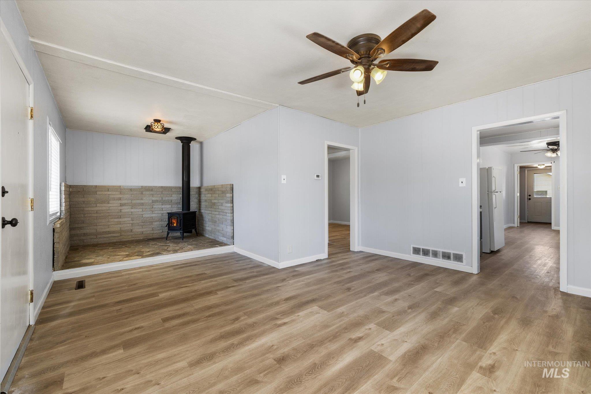 Spacious living area featuring wood-finish flooring, a wood-burning stove with stone surround, a ceiling fan with integrated lighting, and light gray wall finishes
