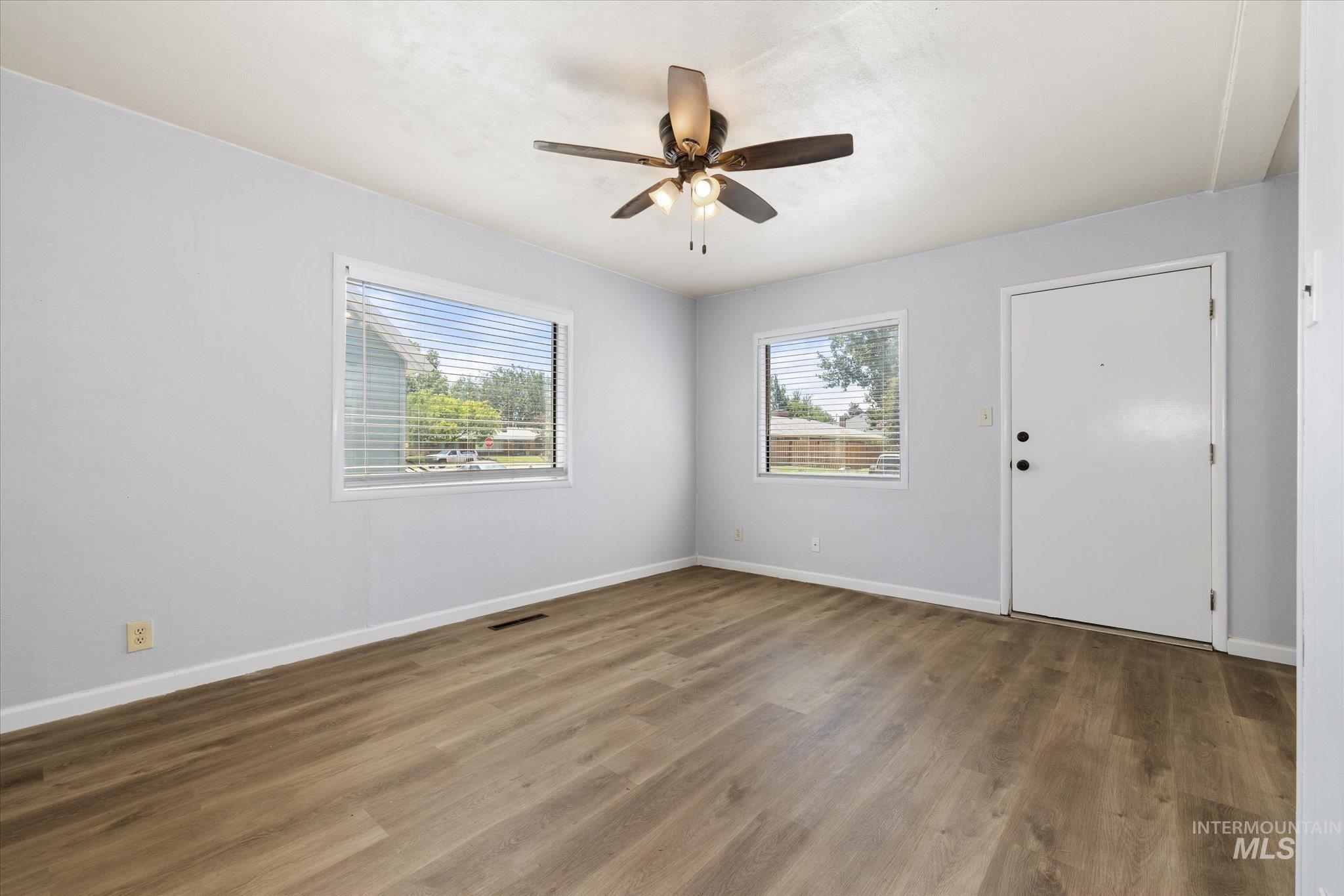 Bright interior space featuring wood-finish flooring, two large windows with blinds, a ceiling fan with integrated lighting, and a white entry door