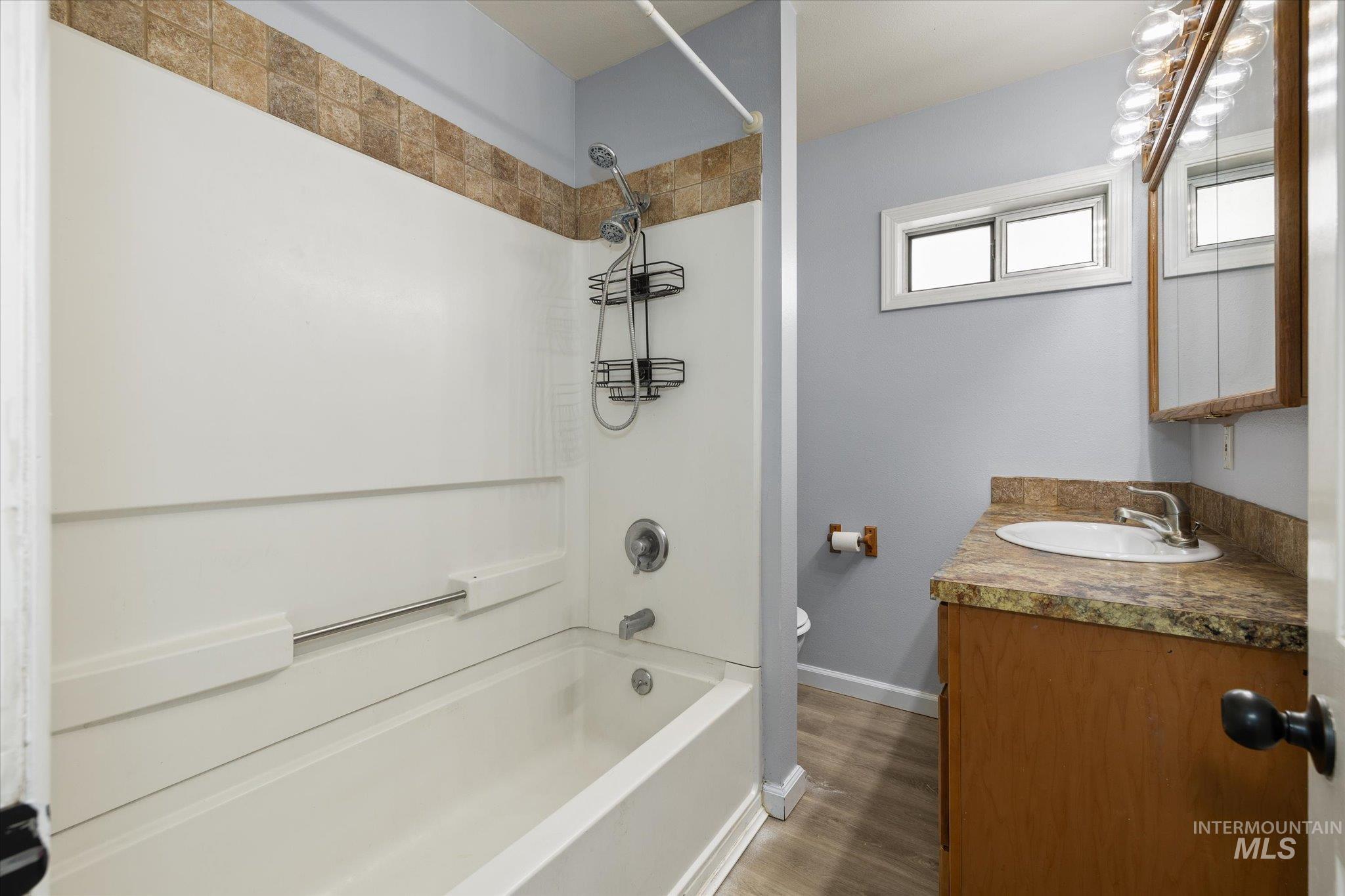 Bathroom featuring a white bathtub with a tiled shower surround, light blue wall paint, a vanity with a stone-finish countertop, a mounted mirror cabinet, and wood-finish flooring