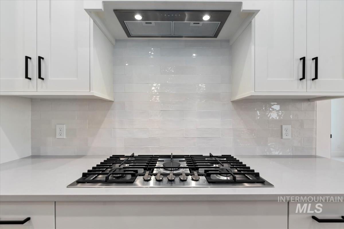 Kitchen view of range hood, stainless steel gas cooktop, white cabinets, decorative backsplash, and light stone counters