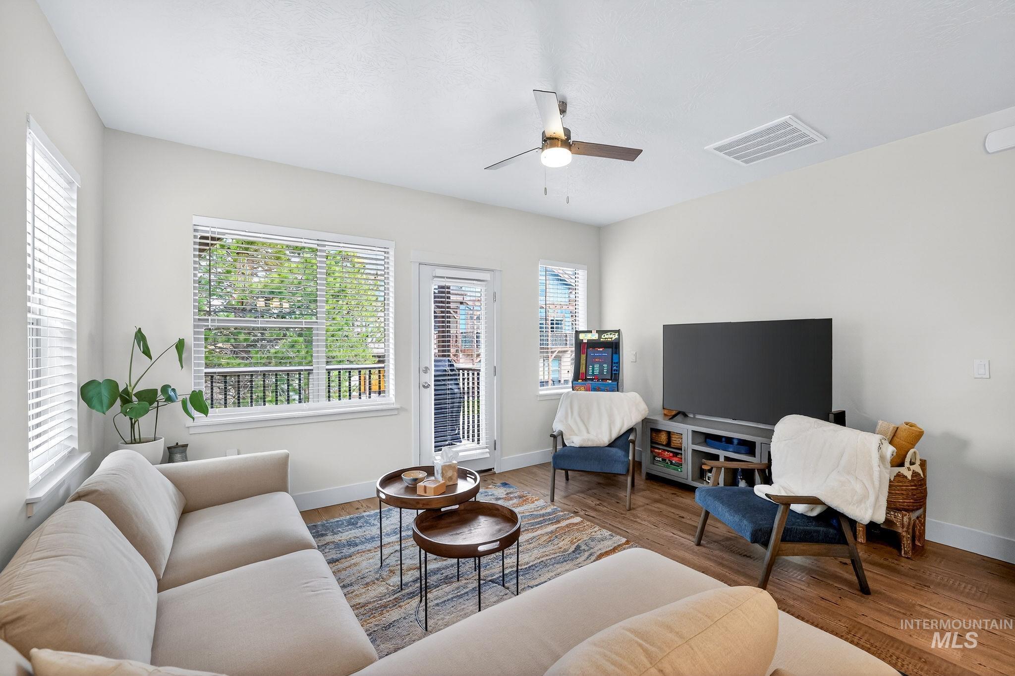 Open-concept living area featuring wood-finish flooring, abundant natural light from multiple windows, a ceiling fan with integrated lighting, and a glass-paneled door leading to an exterior balcony with black iron railings