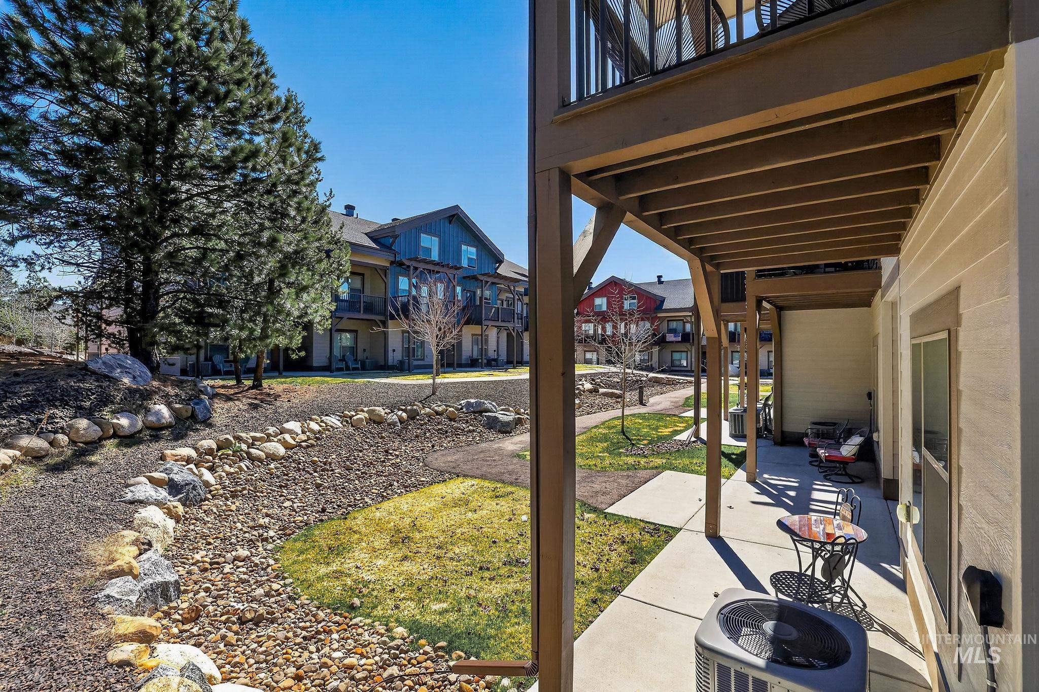 Ground-level patio with overhead balcony, concrete flooring, and direct access to an outdoor area with a decorative rock feature and mature trees