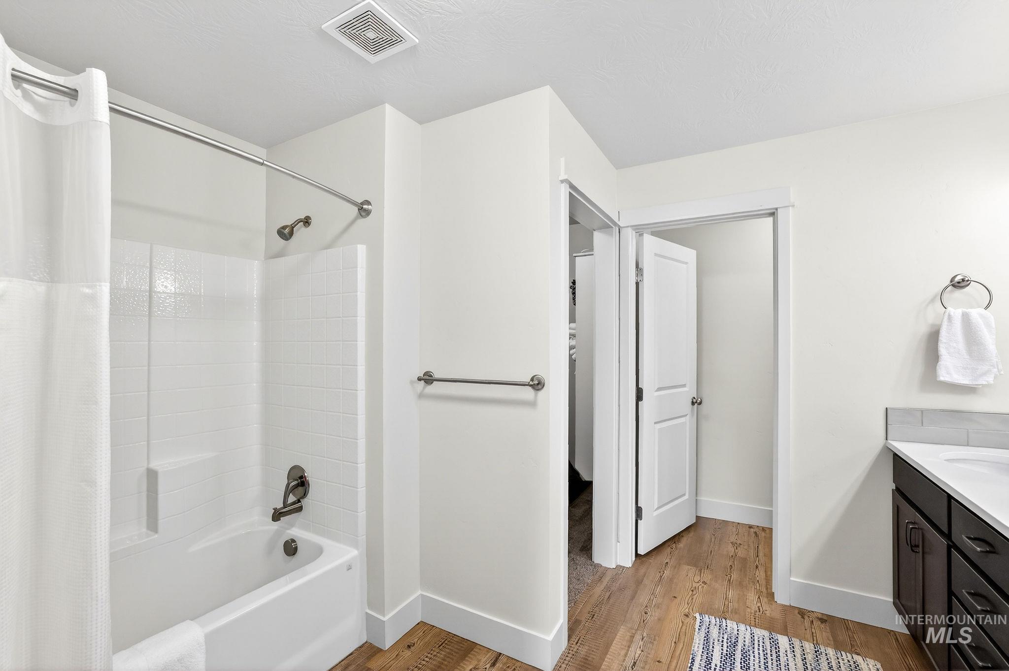 Bathroom featuring a white tub with shower, subway tile surround, wood-finish flooring, and a vanity with dark cabinetry