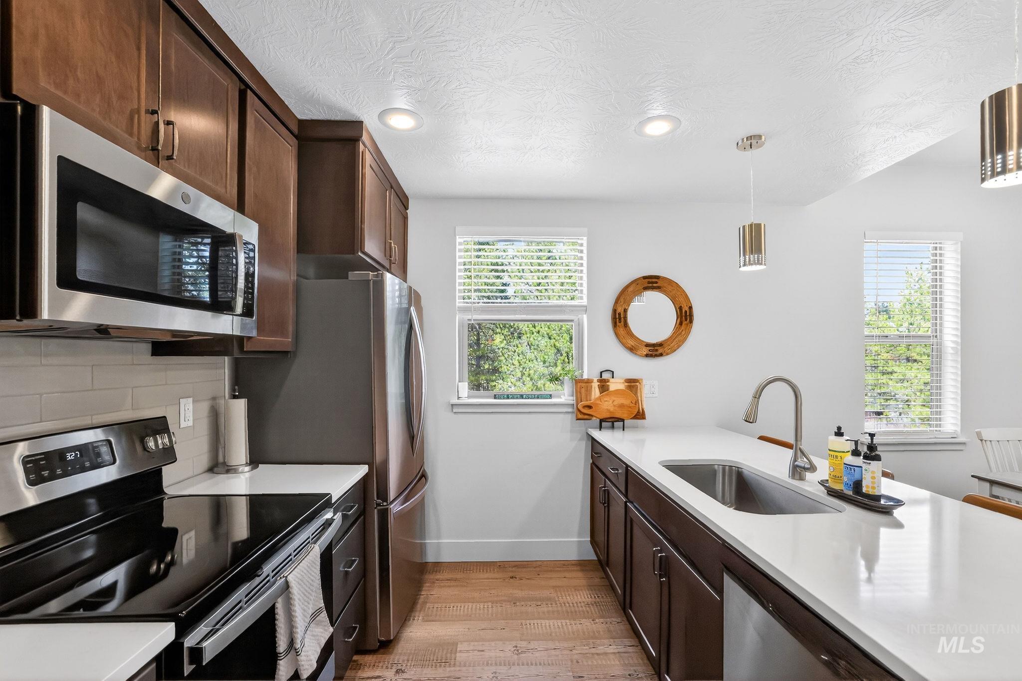 Contemporary kitchen featuring stainless steel appliances, dark wood cabinetry, light-toned countertops, wood-finish flooring, and two windows with blinds