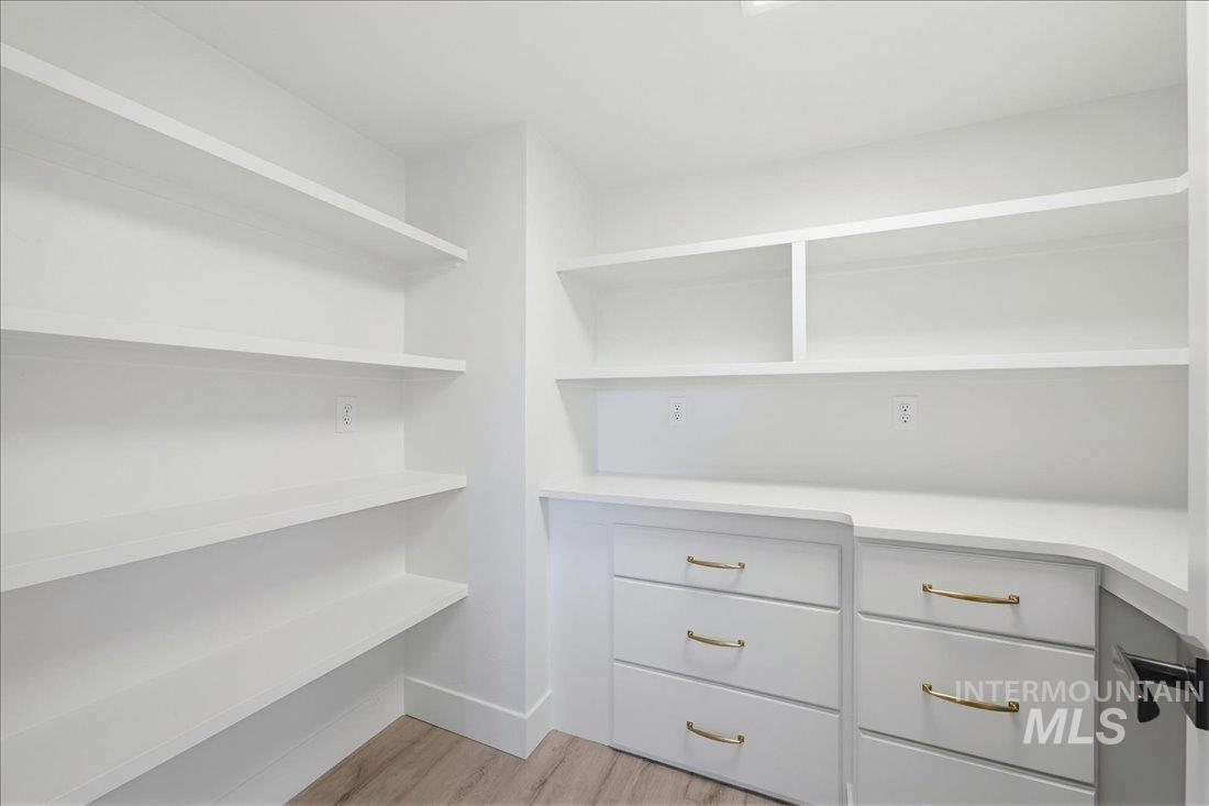 Custom-built pantry featuring extensive white shelving, built-in desk with white countertop, white cabinetry with gold-tone hardware, and wood-finish flooring