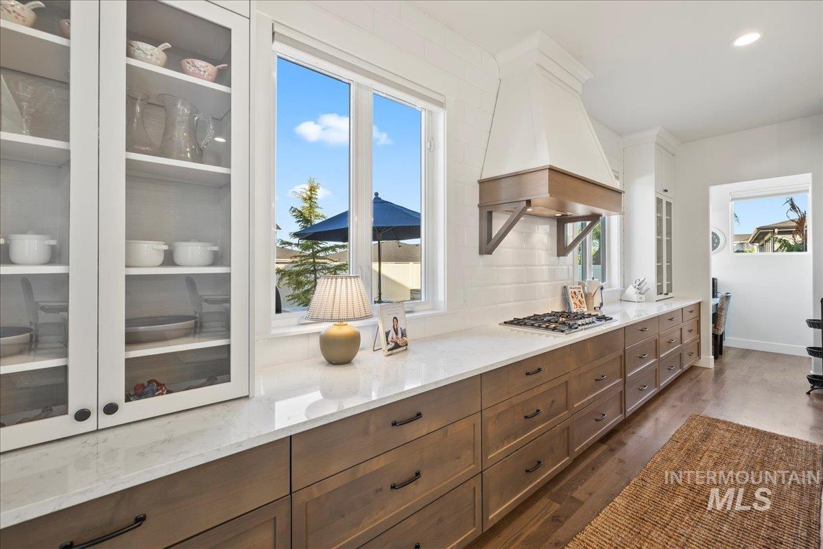 Kitchen with premium range hood, glass insert cabinets, dark wood-type flooring, light stone counters, and backsplash
