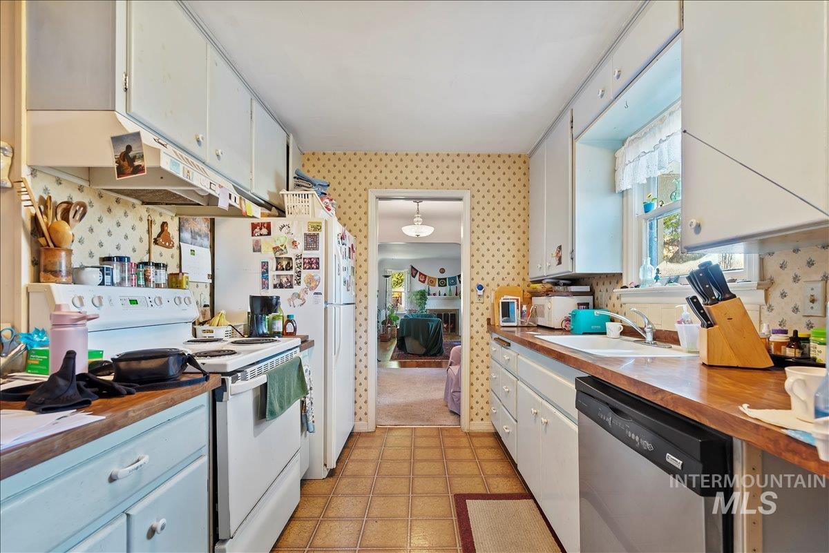 Kitchen with white appliances, wallpapered walls, white cabinets, under cabinet range hood, and pendant lighting