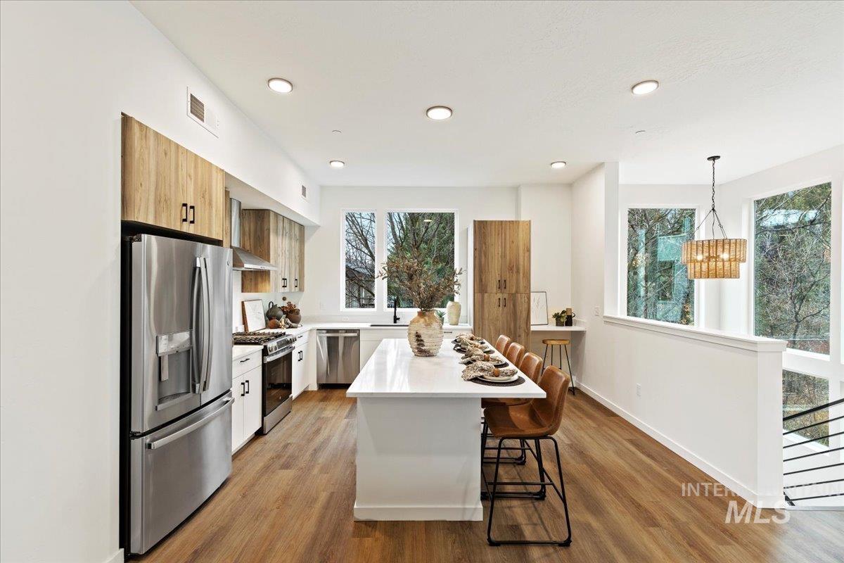 Kitchen featuring stainless steel appliances, a breakfast bar area, light wood finished floors, pendant lighting, and wall chimney range hood