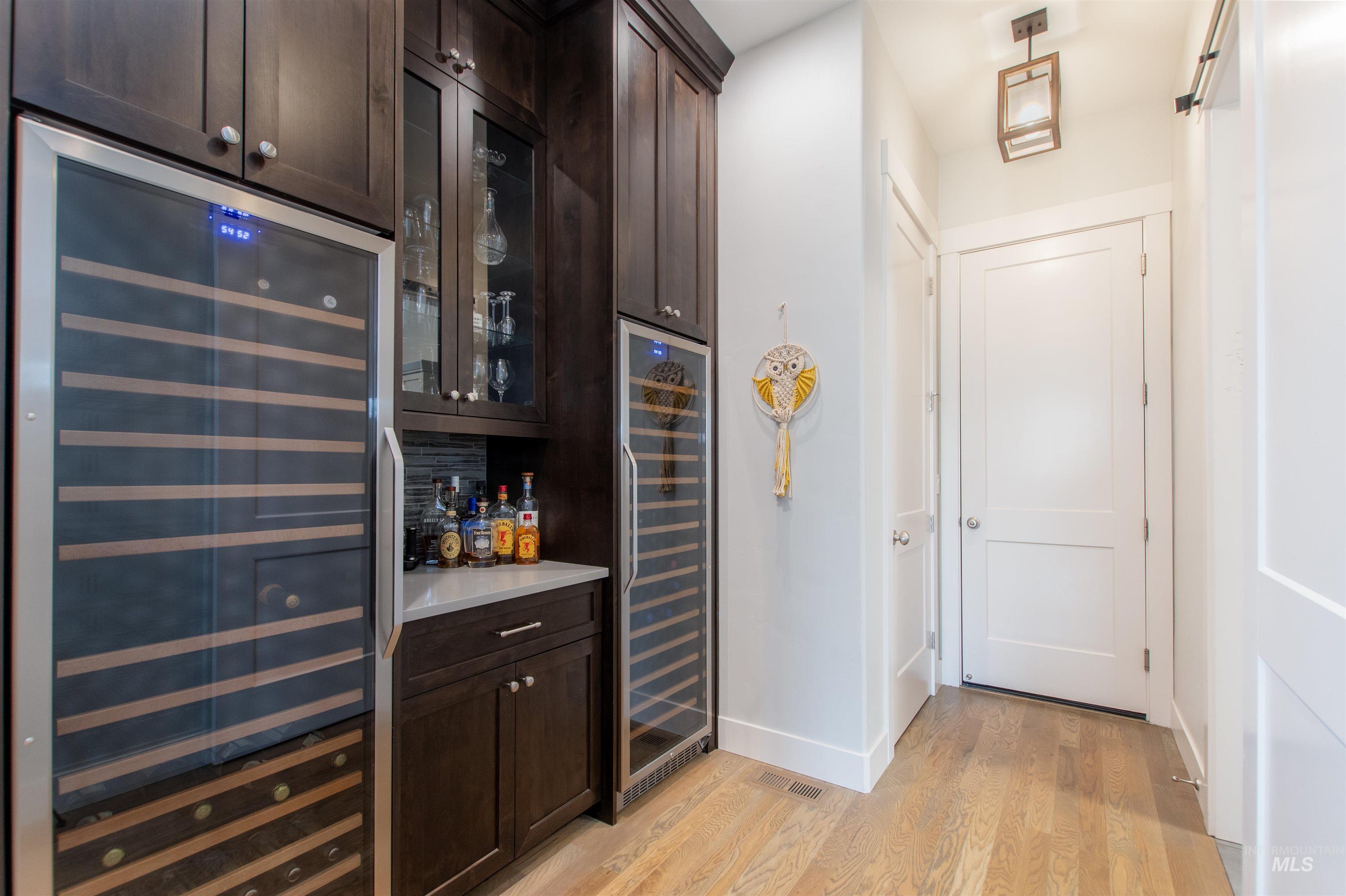 Indoor dry bar with dark brown cabinets, beverage cooler, light wood finished floors, and glass insert cabinets