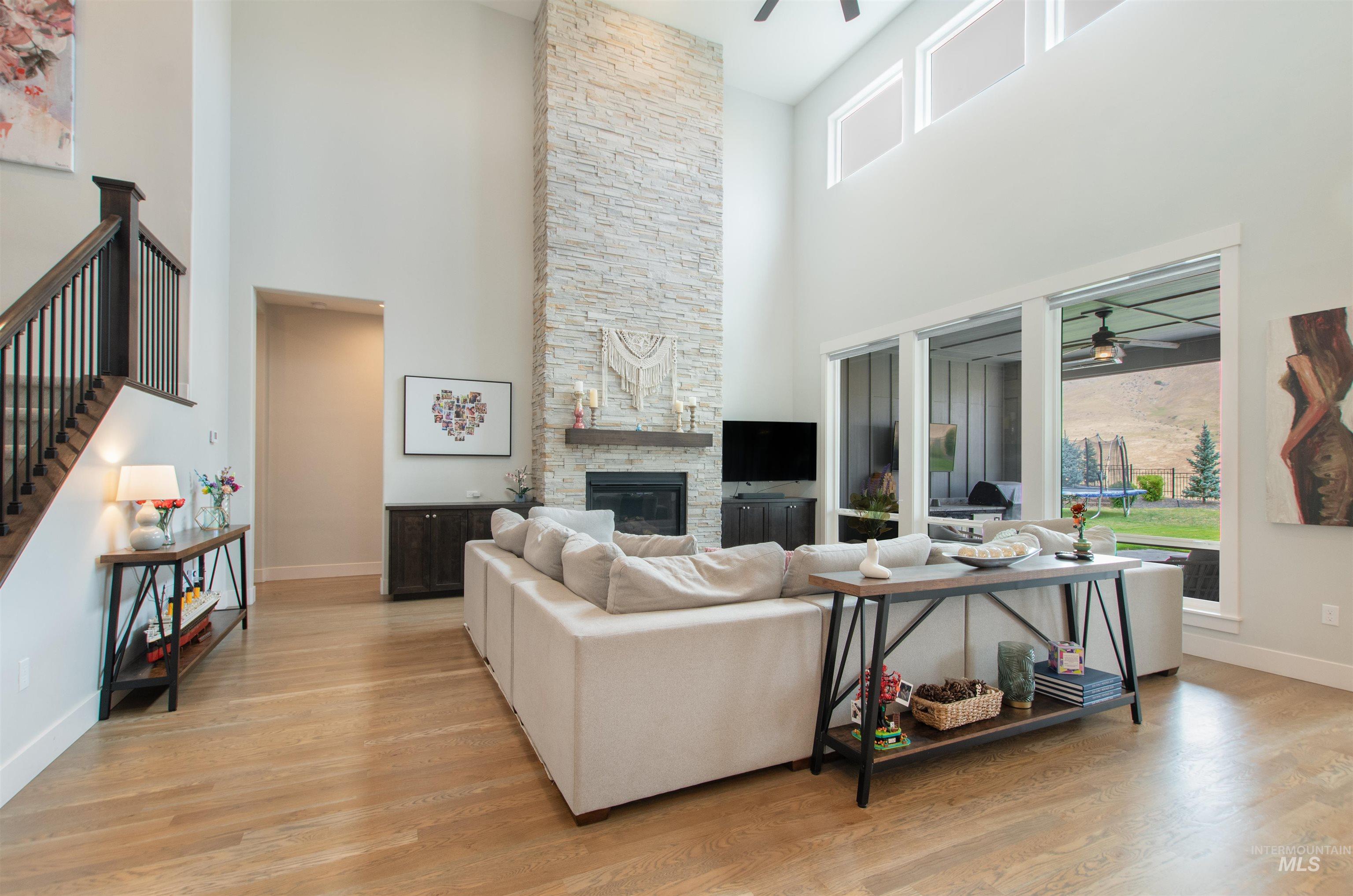 Living area with a ceiling fan, a stone fireplace, light wood-style floors, stairway, and a towering ceiling