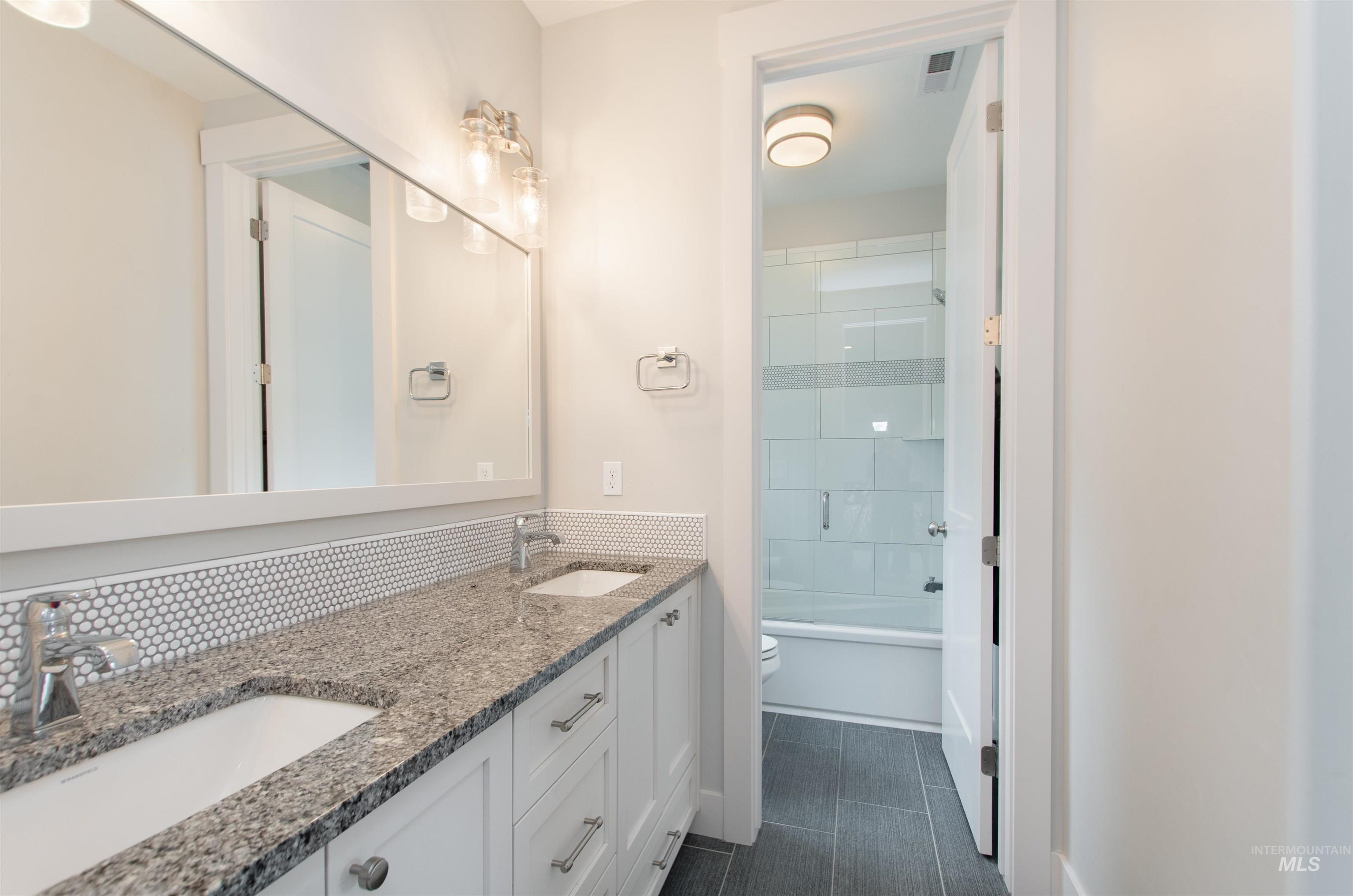 Full bathroom with double vanity, shower / bath combination with glass door, tasteful backsplash, and dark tile patterned flooring