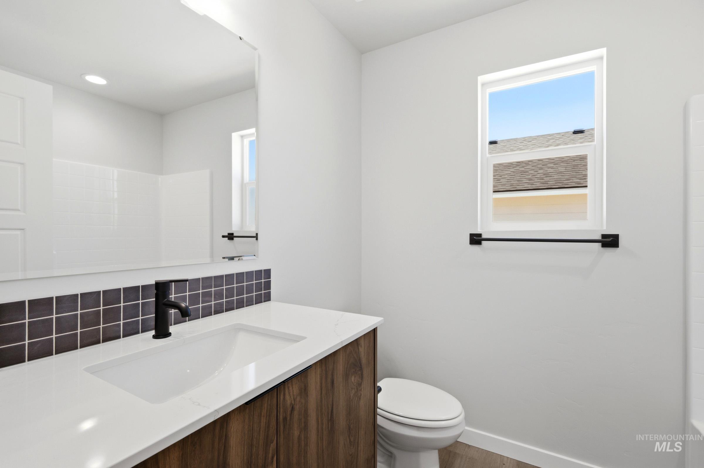 Bathroom vanity featuring a wood-finish cabinet, white countertop with integrated sink, matte black faucet, and a black square tile backsplash