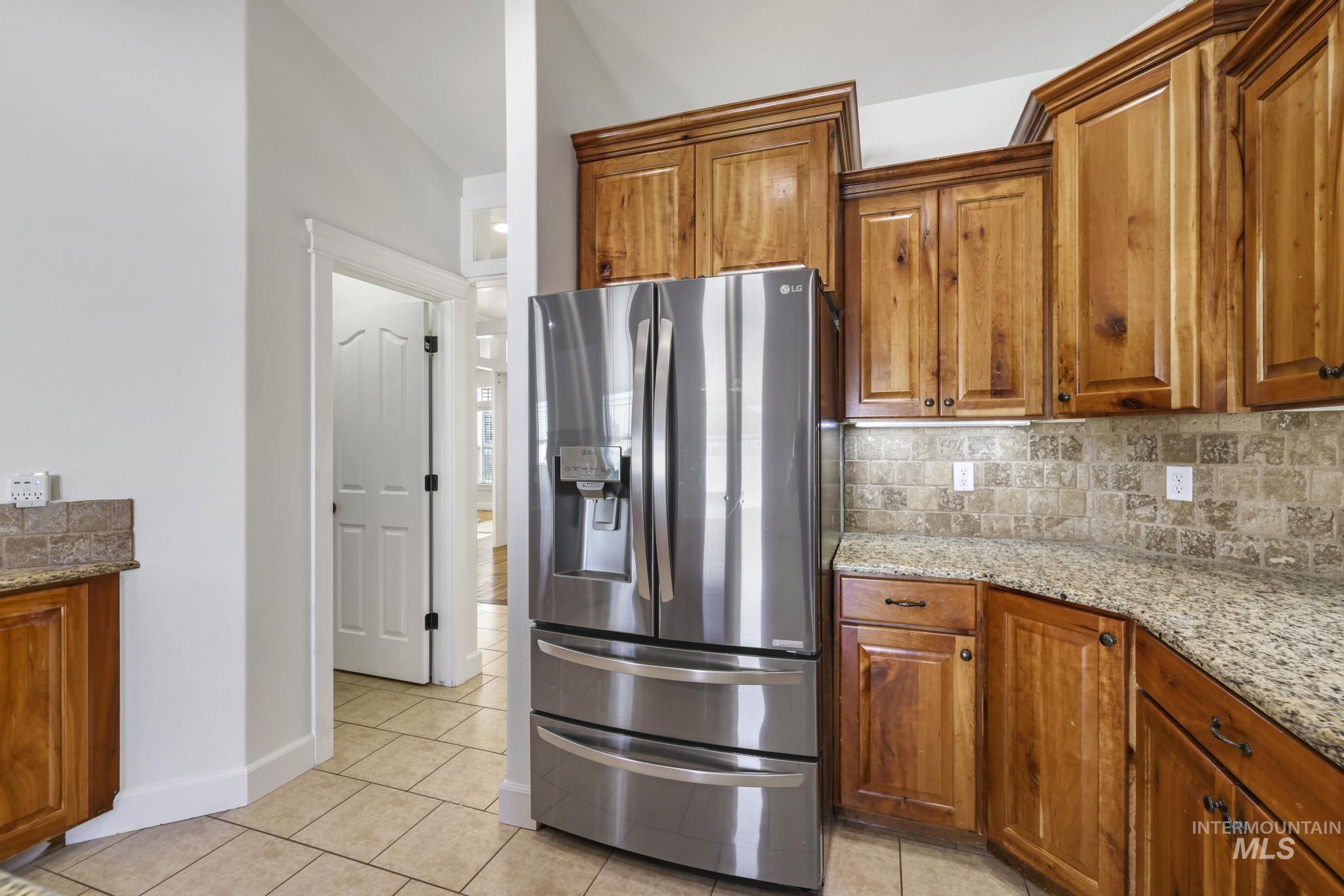 Kitchen with stainless steel fridge with ice dispenser, brown cabinetry, tasteful backsplash, light stone counters, and light tile patterned floors