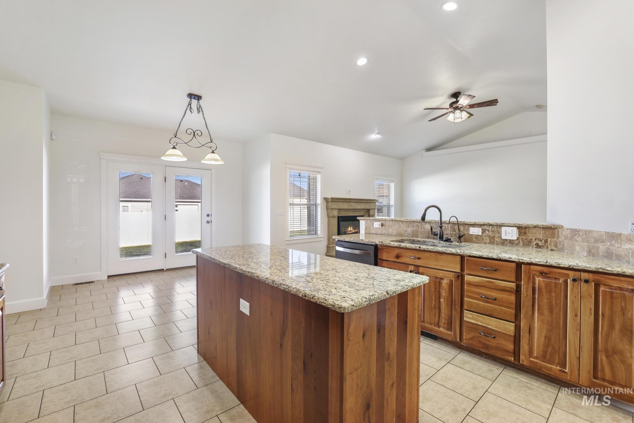 Kitchen with pendant lighting, a lit fireplace, brown cabinetry, light stone counters, and lofted ceiling