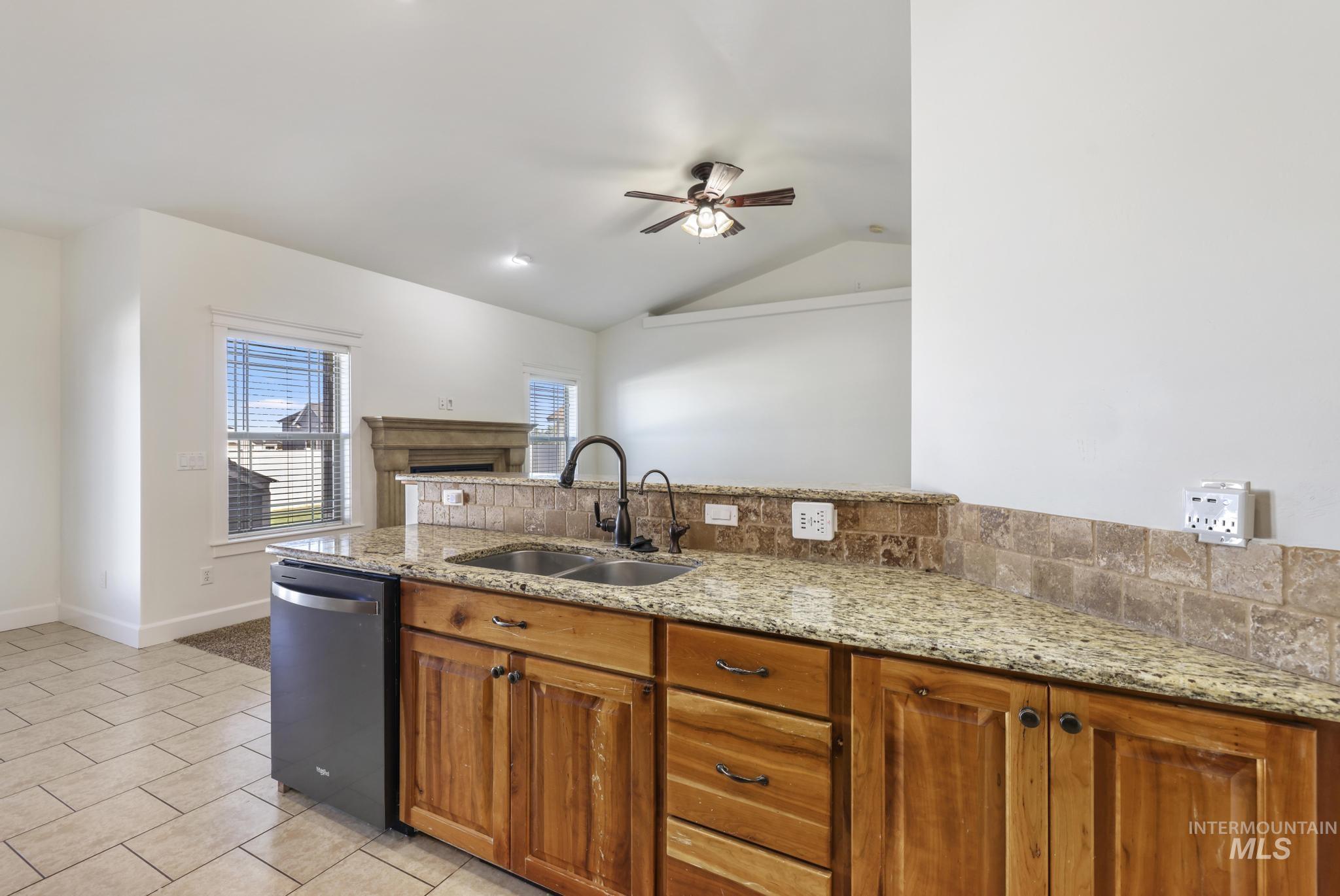 Kitchen with brown cabinets, ceiling fan, vaulted ceiling, stainless steel dishwasher, and light stone countertops