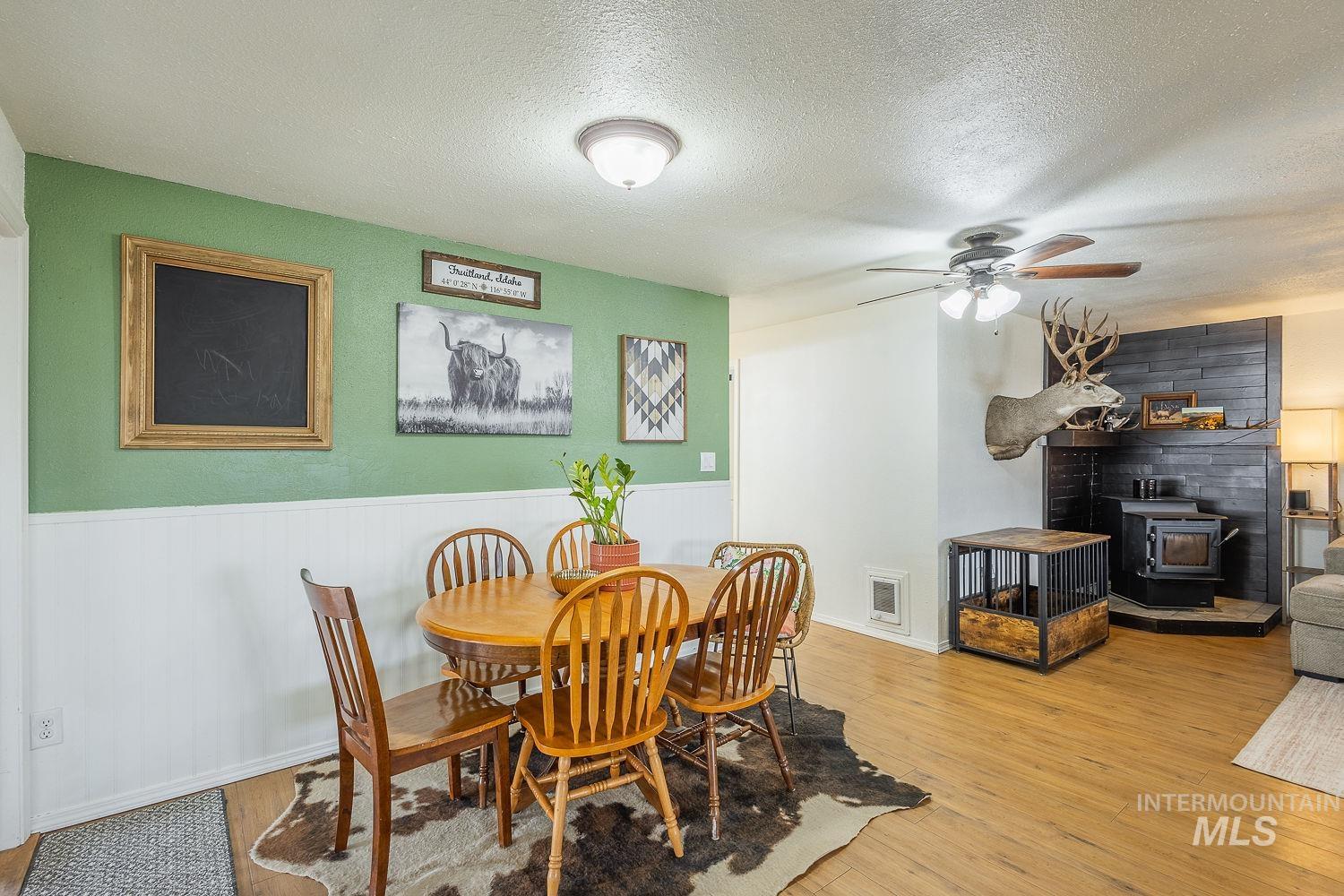 Dining room with a wood stove, a textured ceiling, a ceiling fan, light wood-style flooring, and a wainscoted wall