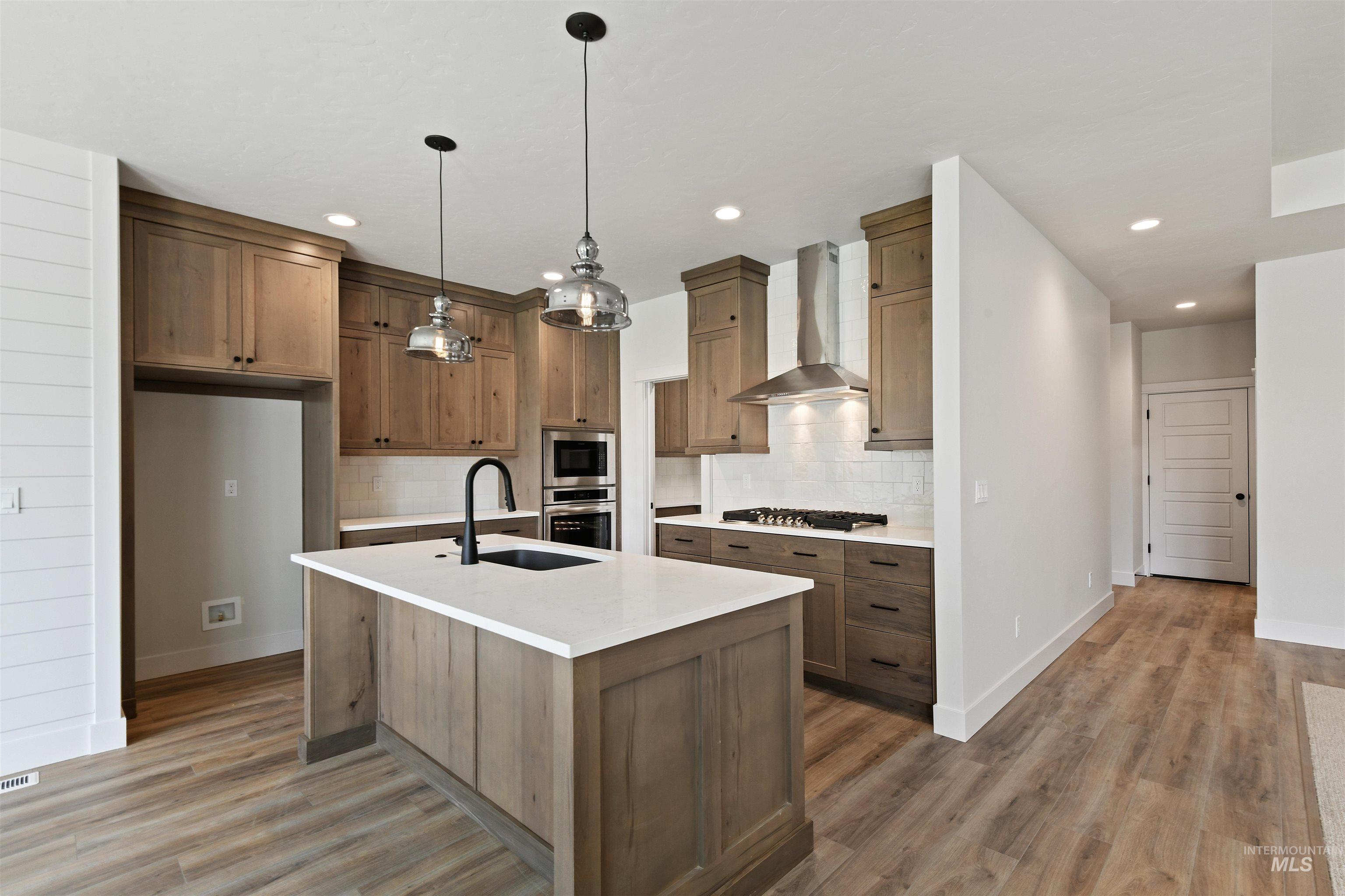 Kitchen featuring wall chimney range hood, stainless steel appliances, tasteful backsplash, light wood-type flooring, and light countertops
