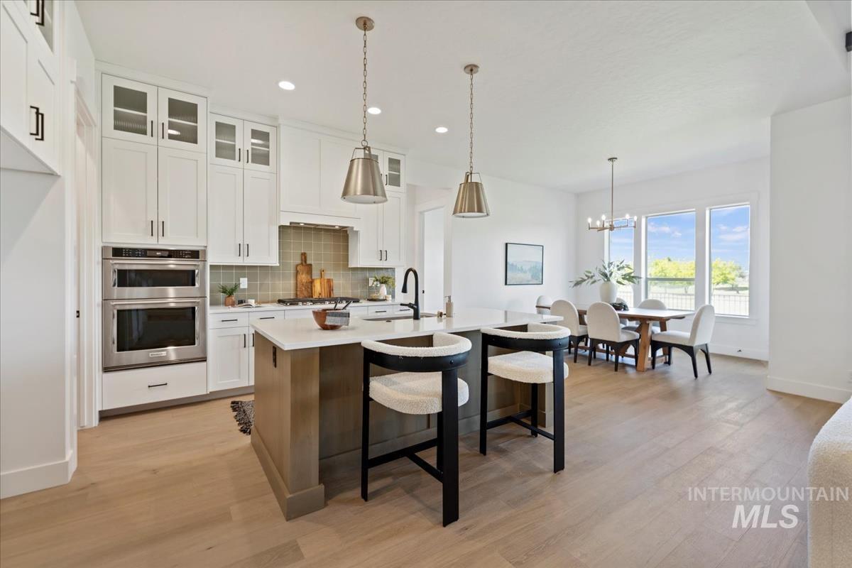 Open-concept kitchen featuring an island with seating, a double wall oven, light wood-finish flooring, white cabinetry, and a dining area with multiple windows