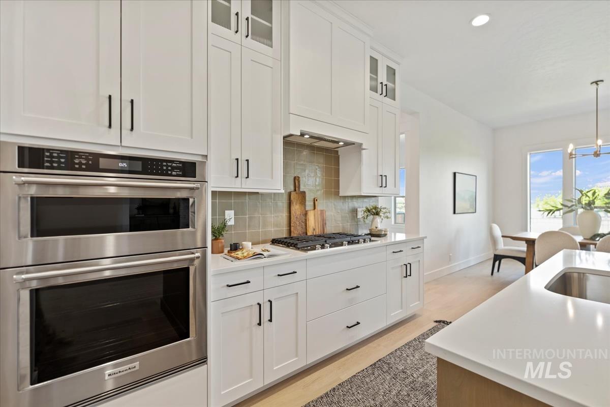 Modern kitchen featuring white shaker cabinetry, stainless steel double ovens, a gas range with a tiled backsplash, wood-finish flooring, and a large kitchen island with an undermount sink