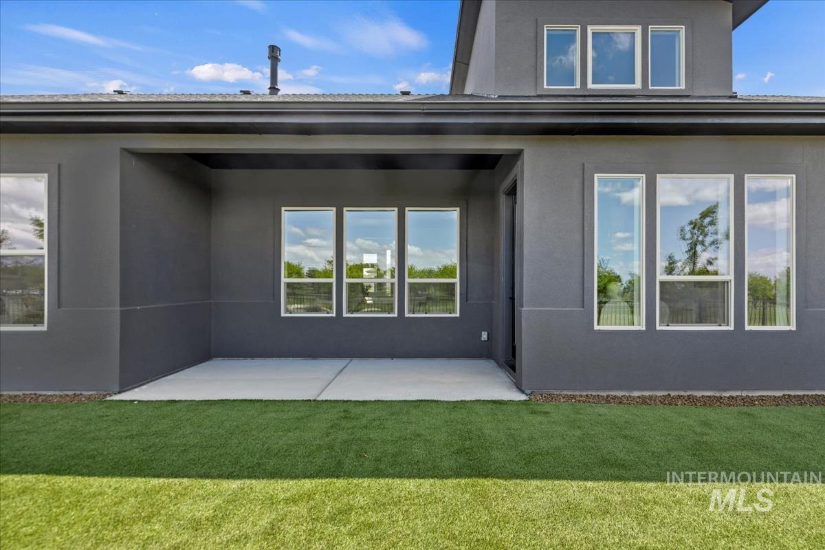 Rear exterior featuring a covered concrete patio, multiple large windows, and a turf lawn