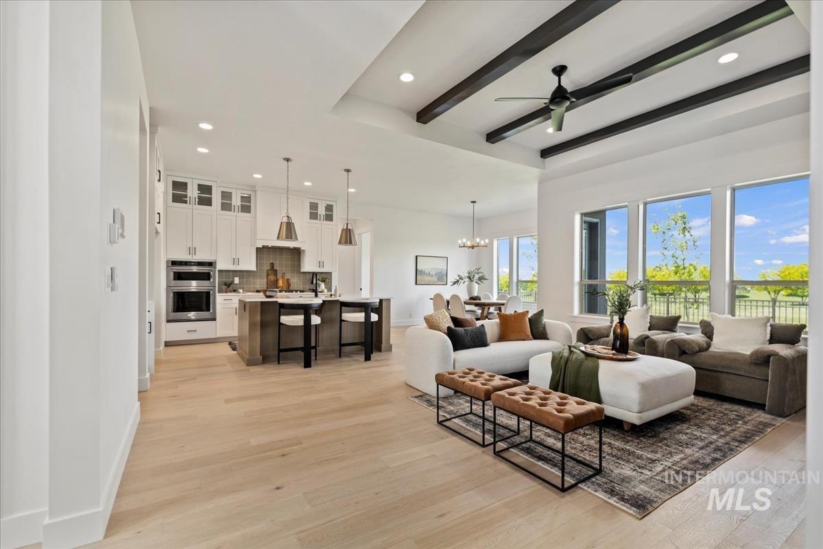 Open-concept living space featuring wood-finish flooring, a kitchen island with seating, a vaulted ceiling with exposed beams and a ceiling fan, and large windows