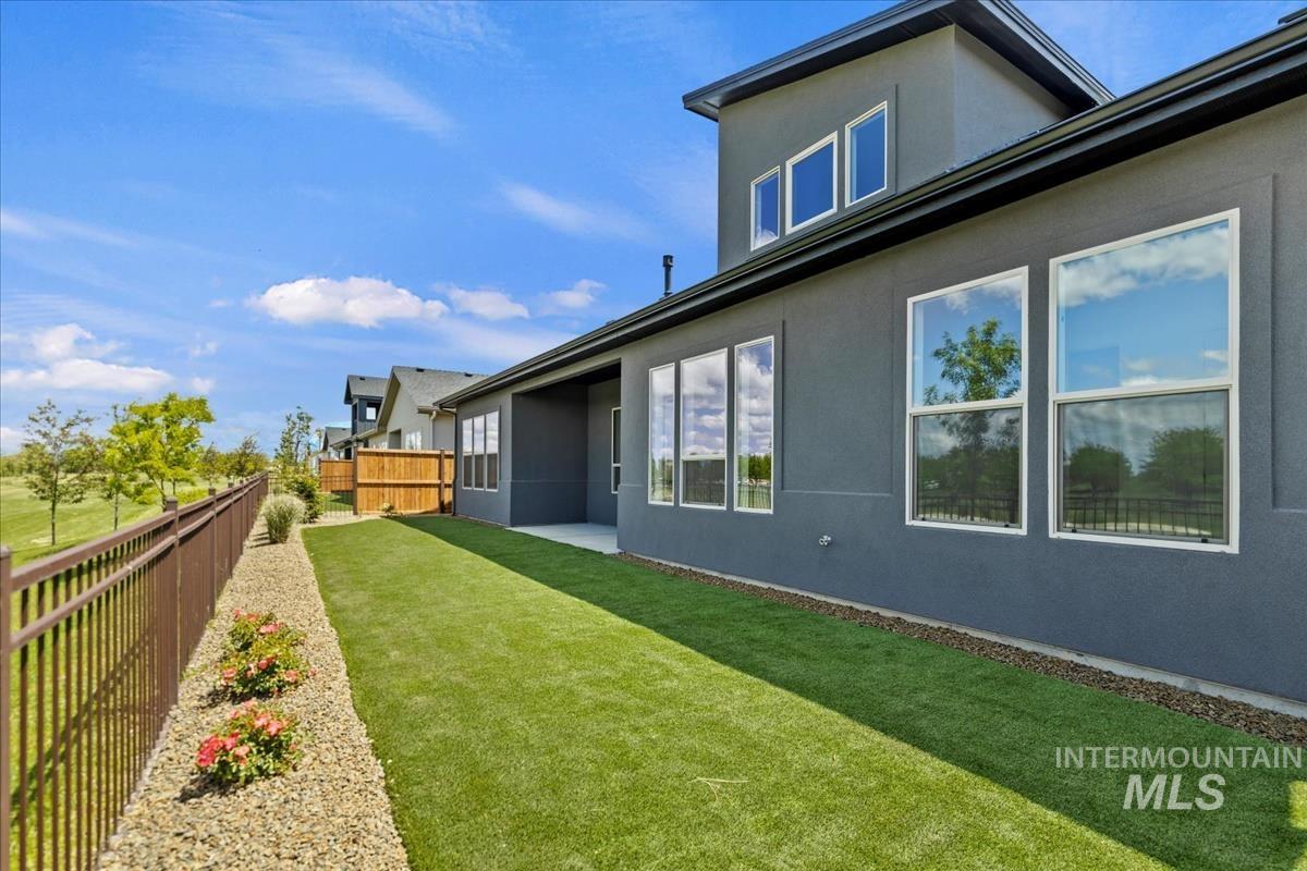 Modern stucco exterior with multiple large windows, a covered patio, and a fenced yard featuring artificial turf and a gravel bed with plantings