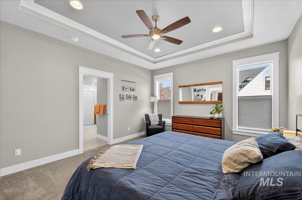 Carpeted bedroom featuring a tray ceiling, multiple windows, ceiling fan, ensuite bath, and recessed lighting