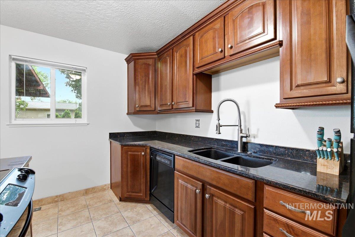 Kitchen featuring wood cabinetry, dark countertops, a double basin sink with a gooseneck faucet, a black dishwasher, and tile flooring