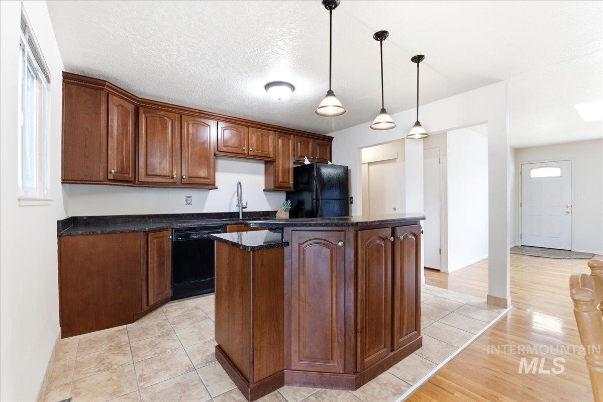 Kitchen featuring wood cabinetry, dark countertops, black appliances, a central island with pendant lighting, and tiled flooring