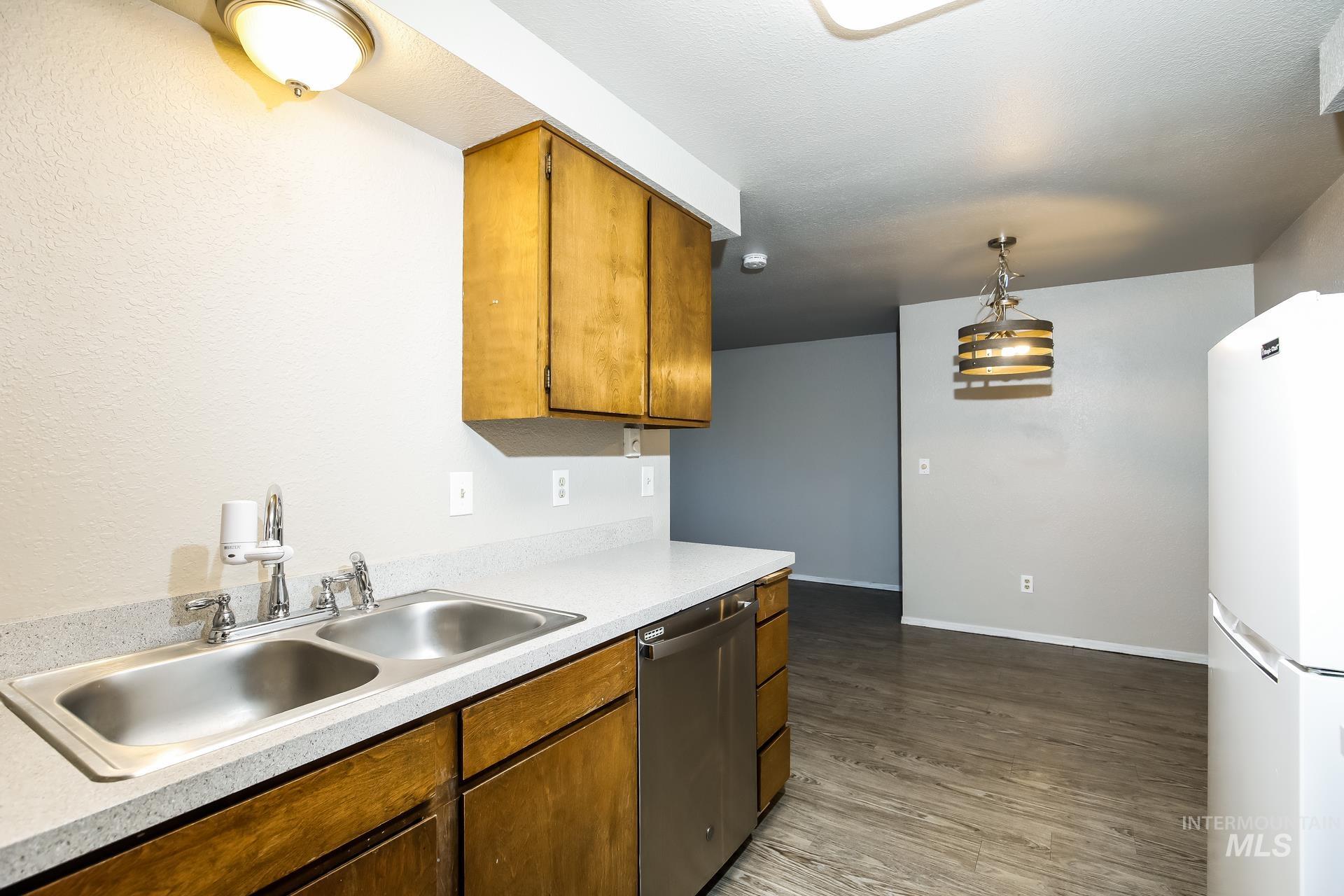 Kitchen featuring freestanding refrigerator, brown cabinetry, light countertops, stainless steel dishwasher, and pendant lighting