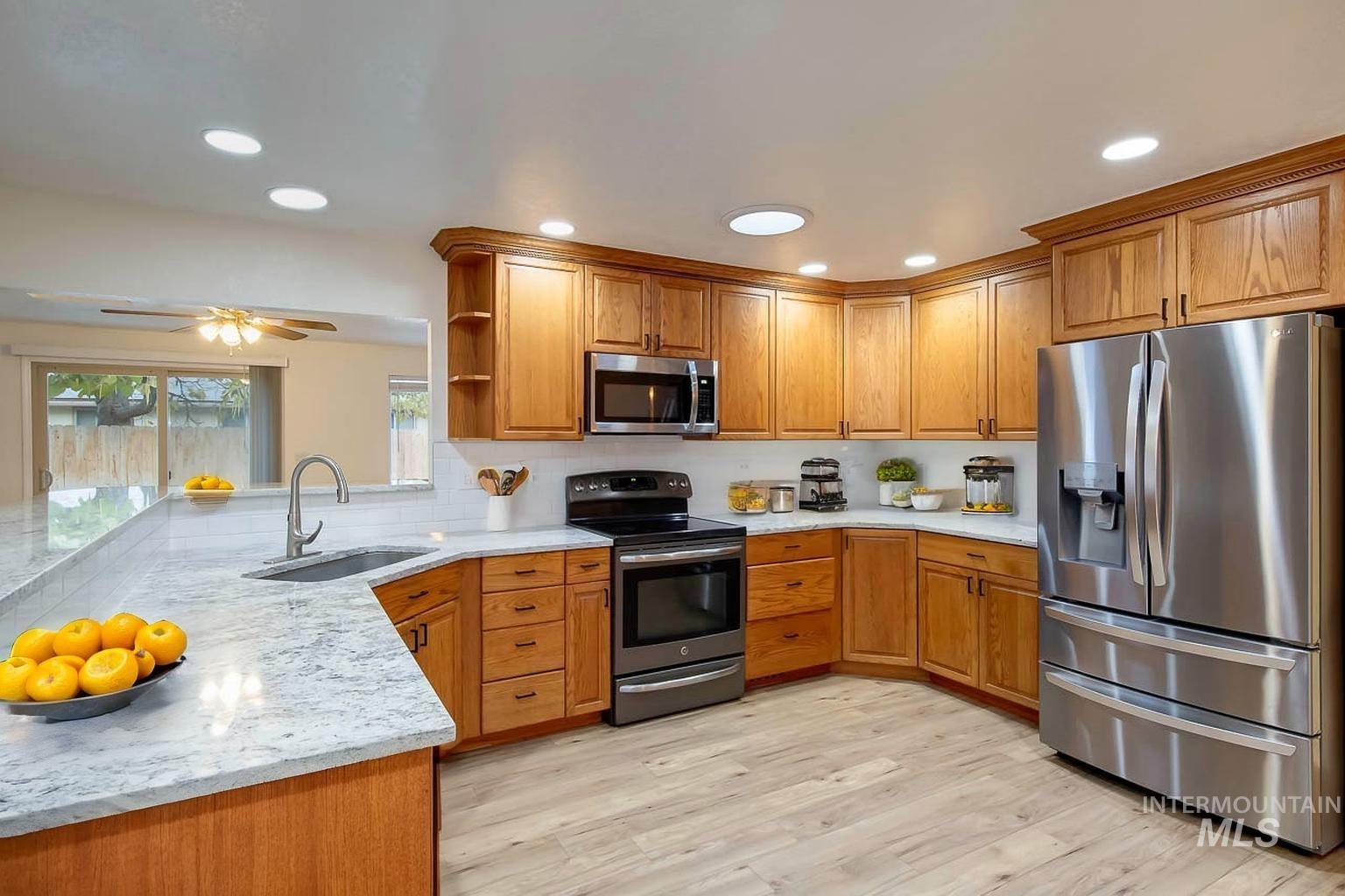 Kitchen featuring stainless steel appliances, light stone countertops, open shelves, brown cabinetry, and ceiling fan