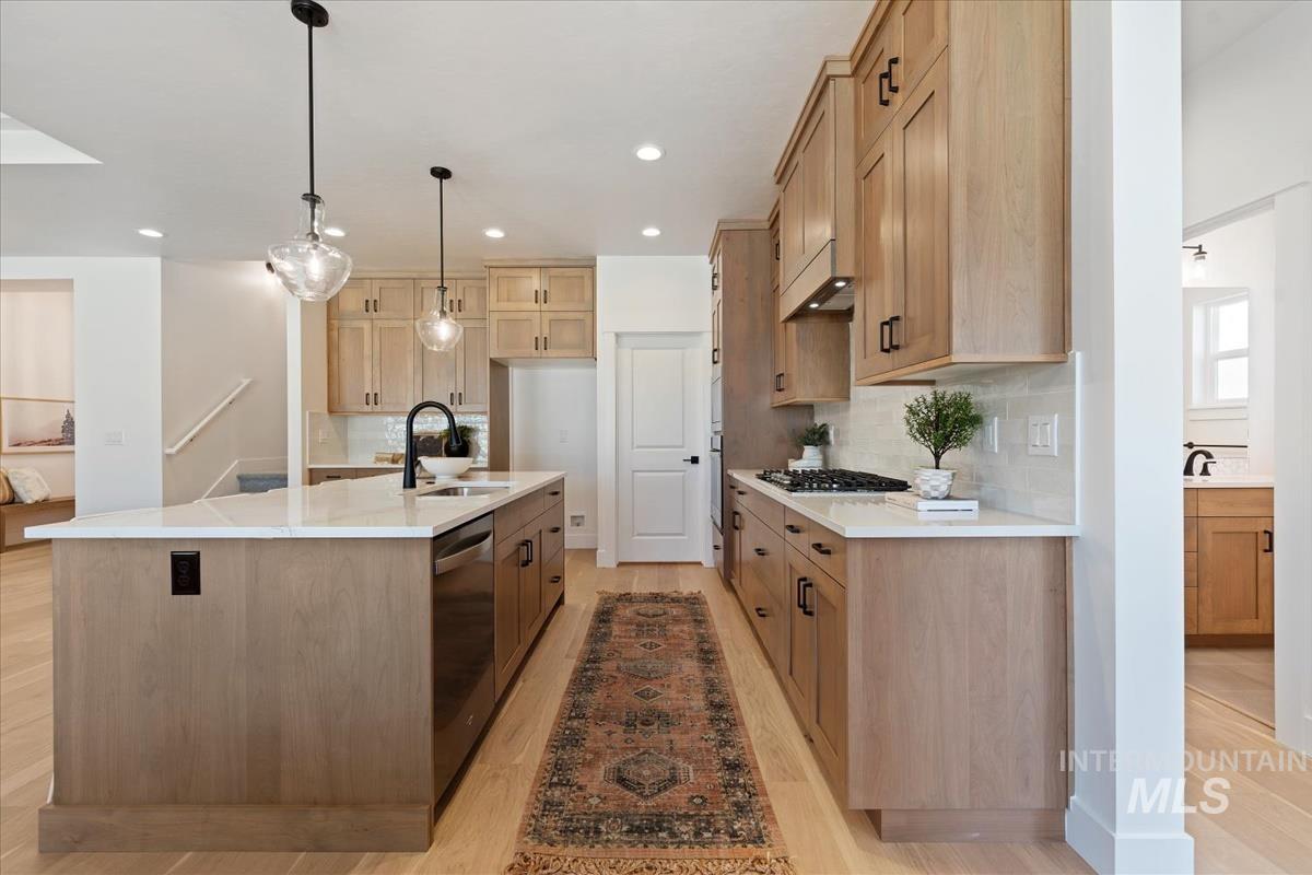 Kitchen featuring light stone countertops, backsplash, light wood-type flooring, hanging light fixtures, and a center island with sink