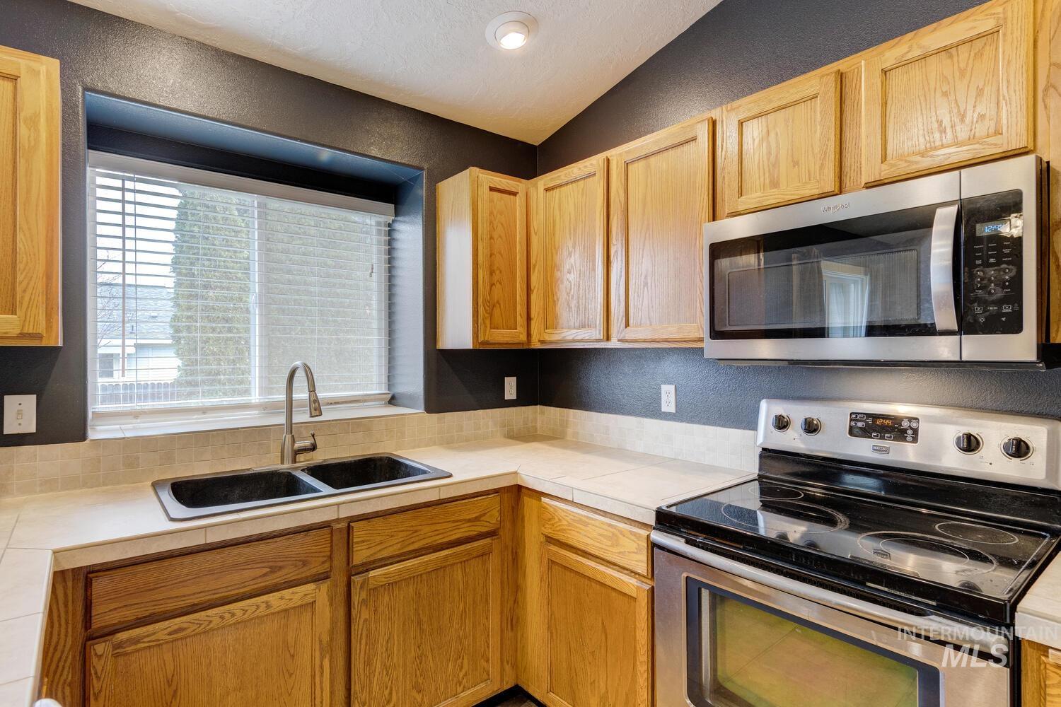 Kitchen with stainless steel appliances, a textured wall, tile countertops, and light brown cabinetry