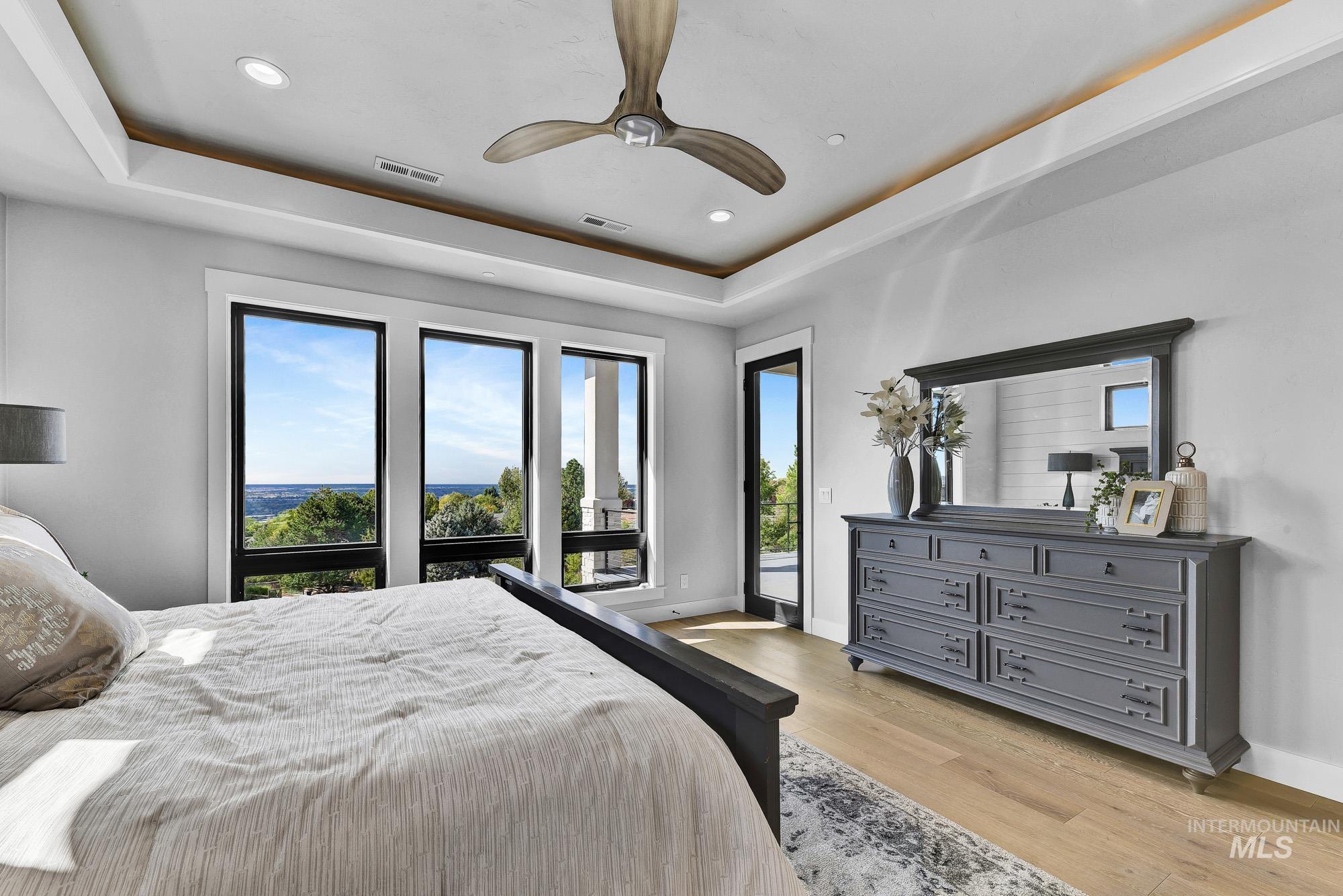Bedroom with a raised ceiling, light wood finished floors, a ceiling fan, recessed lighting, and access to exterior