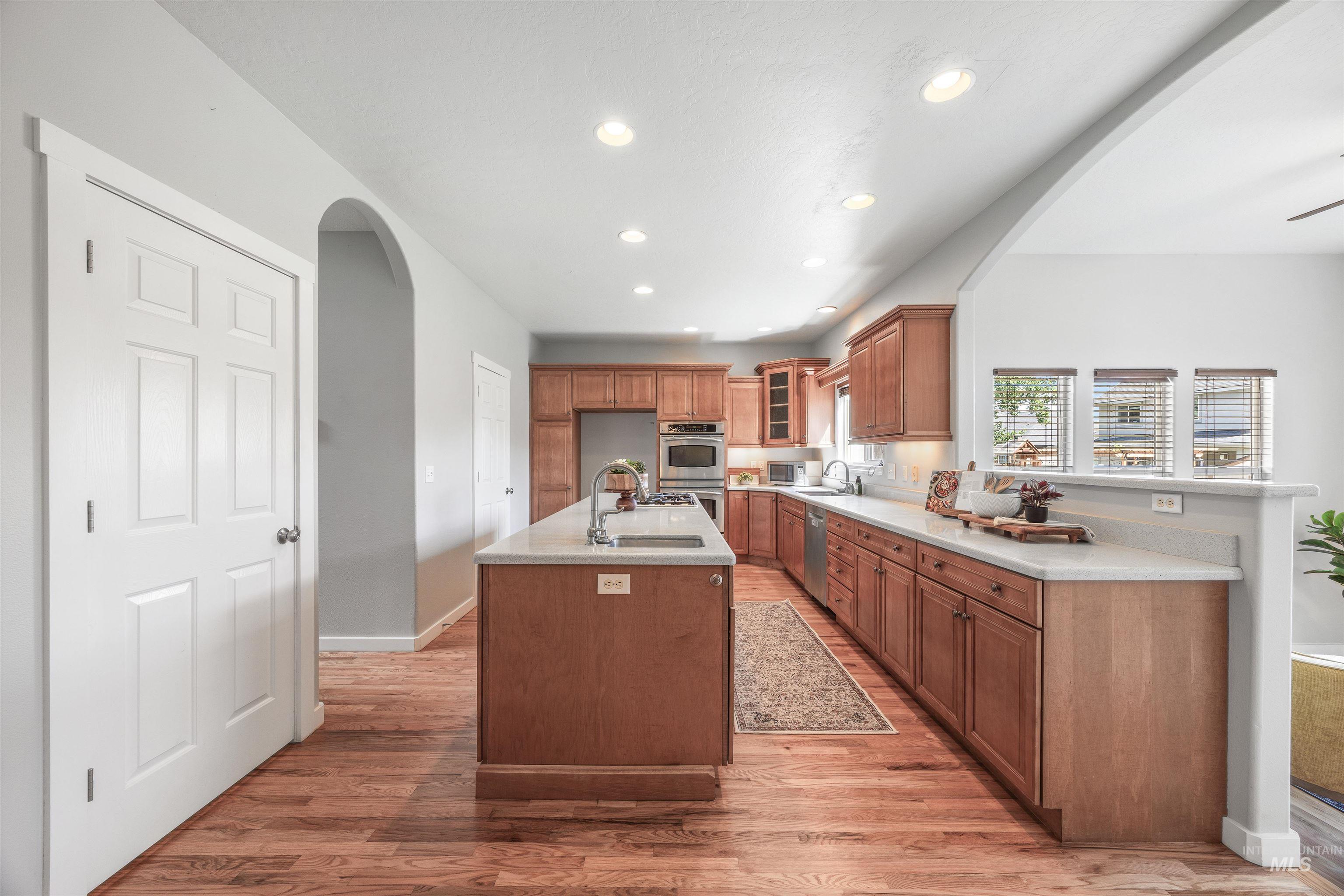 Kitchen with arched walkways, brown cabinets, recessed lighting, glass insert cabinets, and light wood finished floors