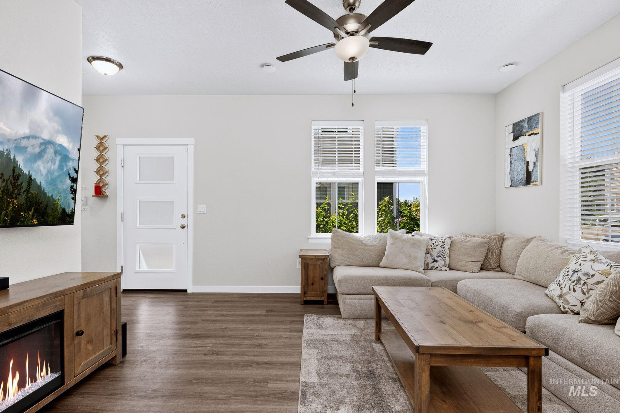 Bright living area with wood-finish flooring, an electric fireplace, multiple windows with blinds, a ceiling fan, and a white paneled door with frosted glass inserts