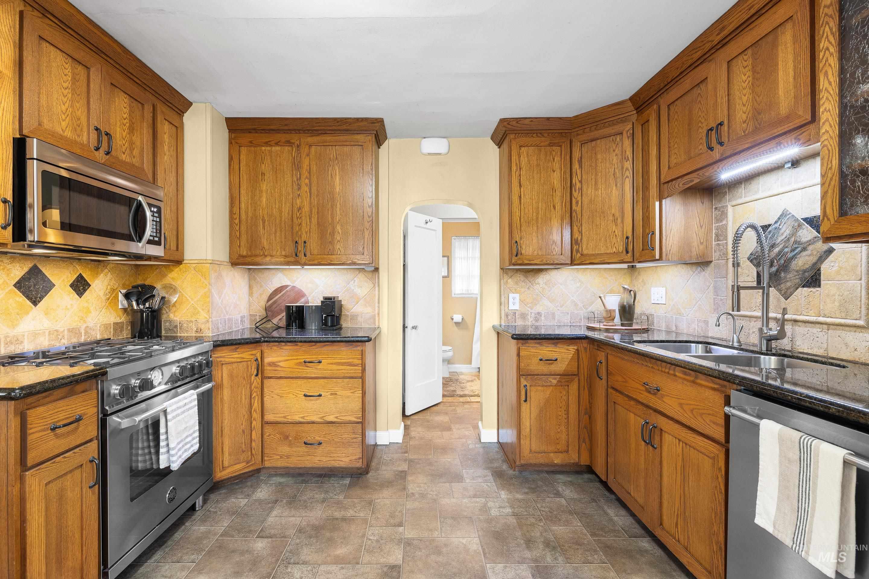 Kitchen with stainless steel appliances, arched walkways, brown cabinetry, dark stone countertops, and backsplash