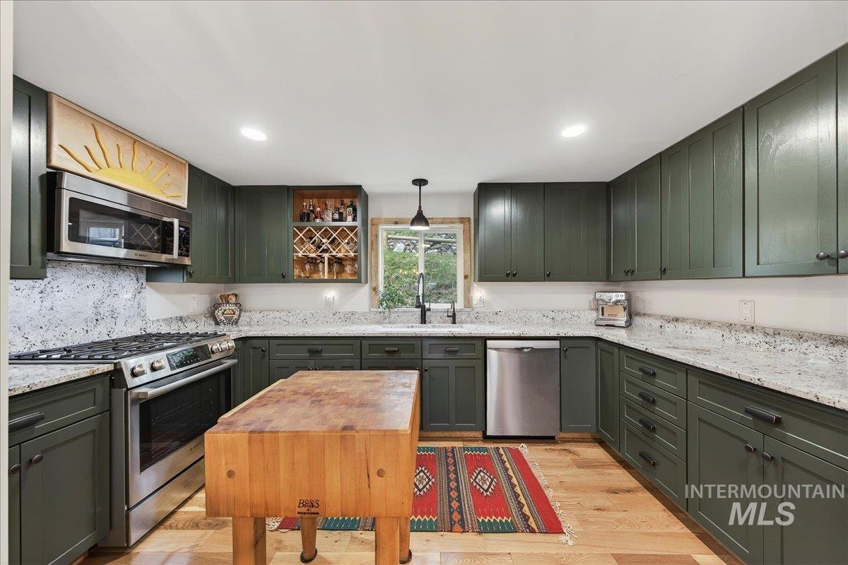 Kitchen featuring green cabinetry, stainless steel appliances, light wood-type flooring, hanging light fixtures, and light stone counters
