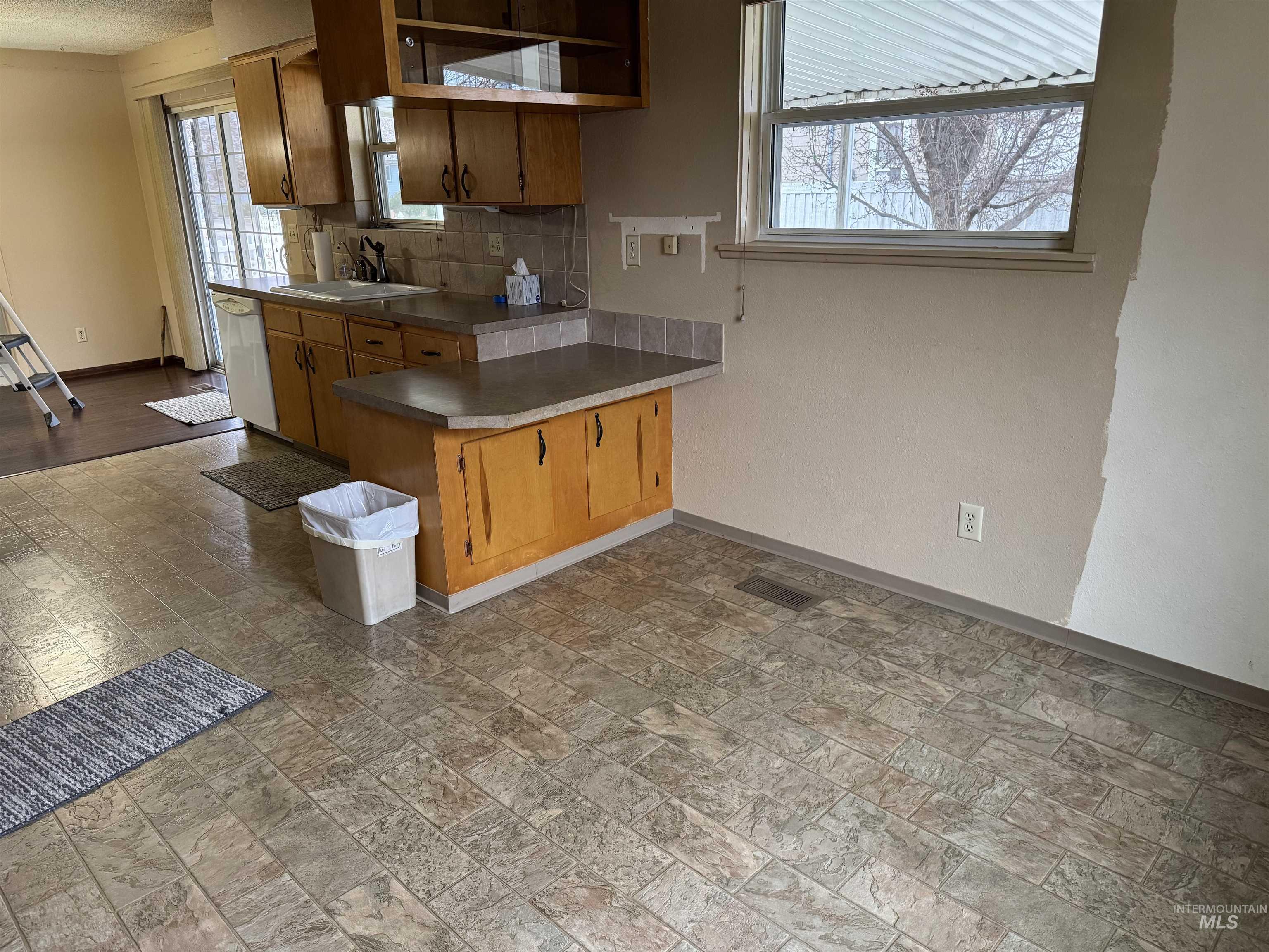 Kitchen with a peninsula, stainless steel dishwasher, brown cabinets, decorative backsplash, and light stone finish floors
