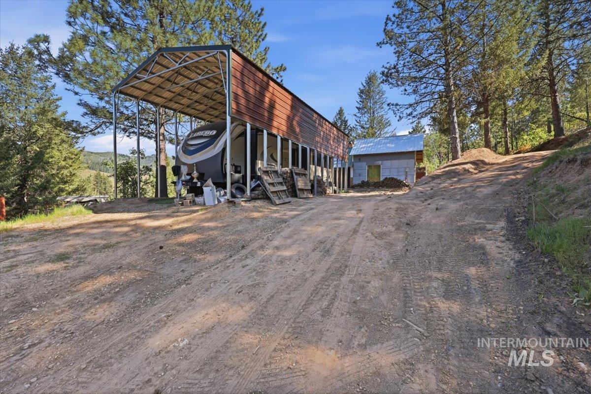 Expansive dirt lot featuring a large metal carport structure with partial wood-finish siding, a separate detached building, and natural wooded surroundings