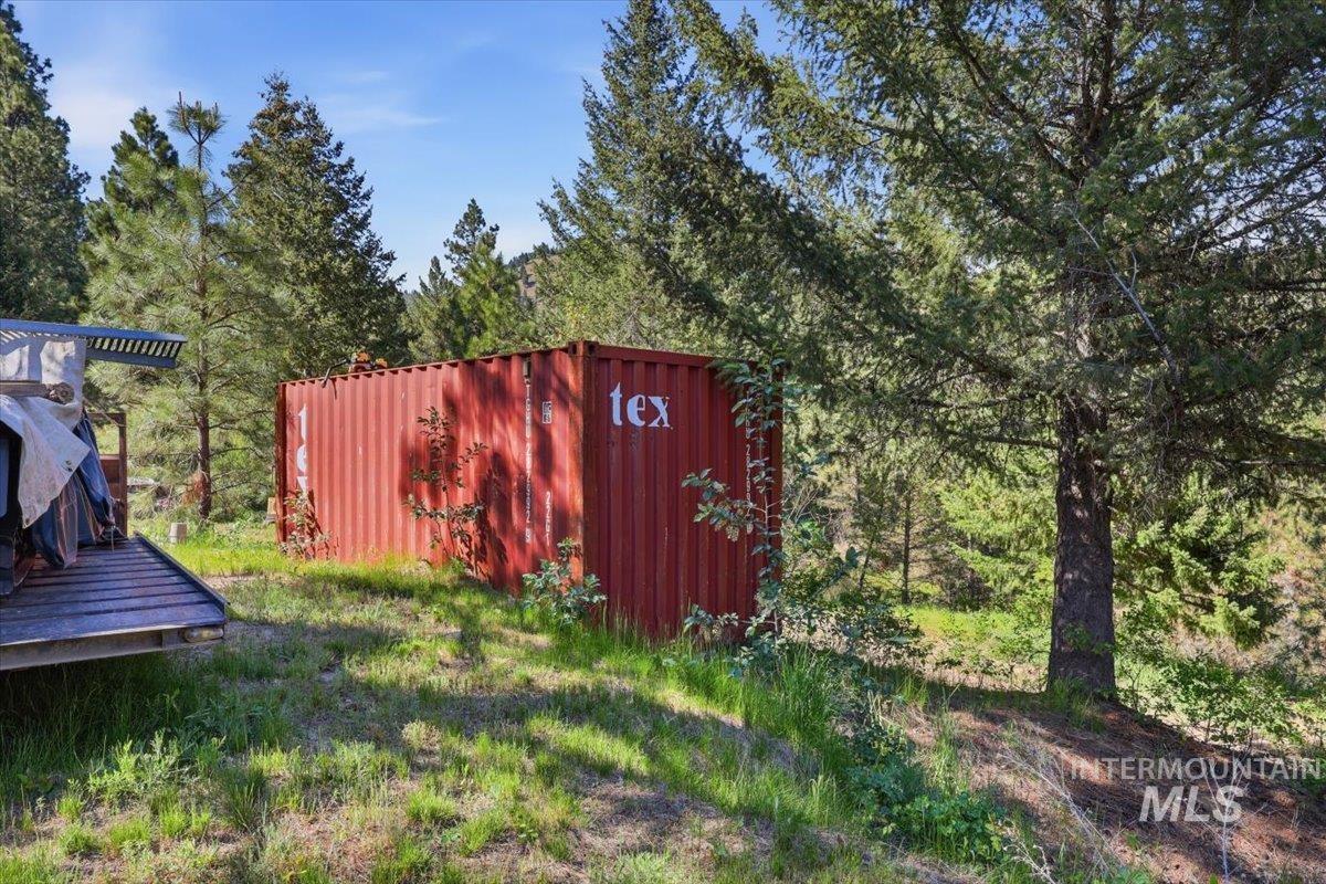 Red shipping container on a sloped, grassy terrain with mature evergreen trees and natural foliage