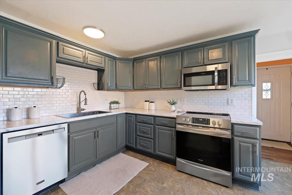 Kitchen with stainless steel appliances, gray cabinets, light stone countertops, and tasteful backsplash