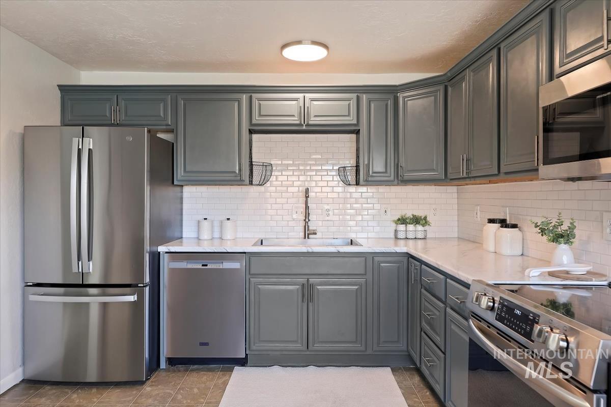 Kitchen featuring stainless steel appliances, gray cabinets, light stone countertops, dark tile patterned flooring, and backsplash