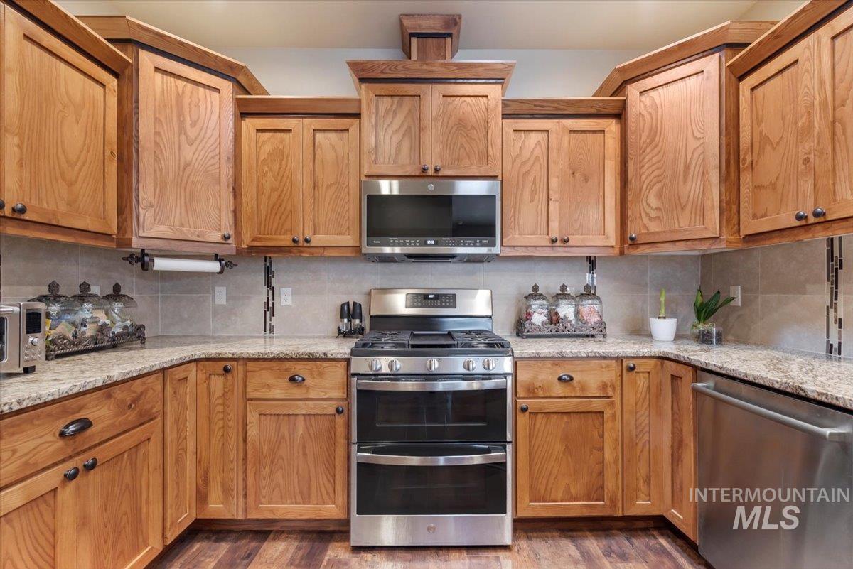 Kitchen featuring stainless steel appliances, backsplash, light stone counters, dark wood-style floors, and brown cabinets