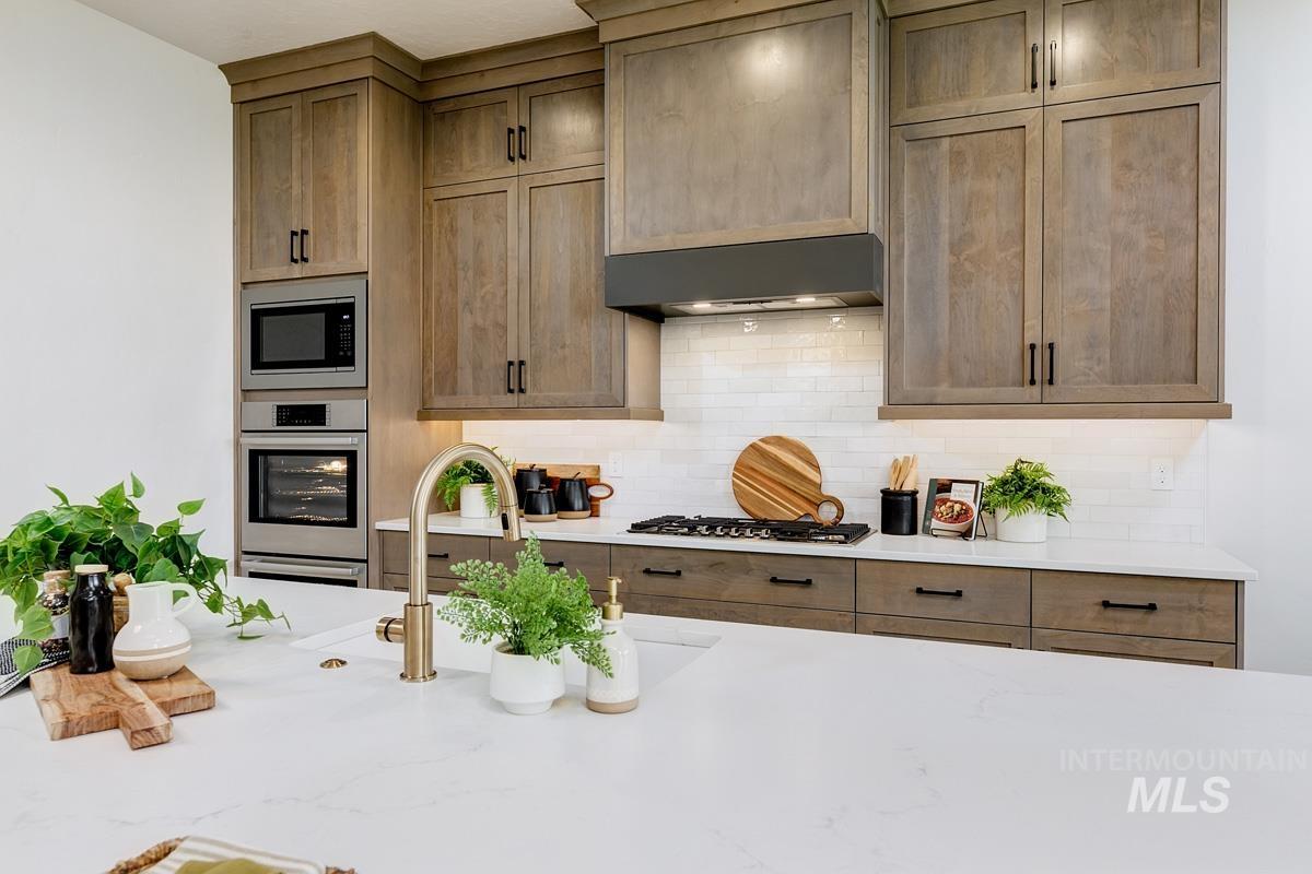 Kitchen with stainless steel appliances, light stone countertops, ventilation hood, decorative backsplash, and brown cabinets